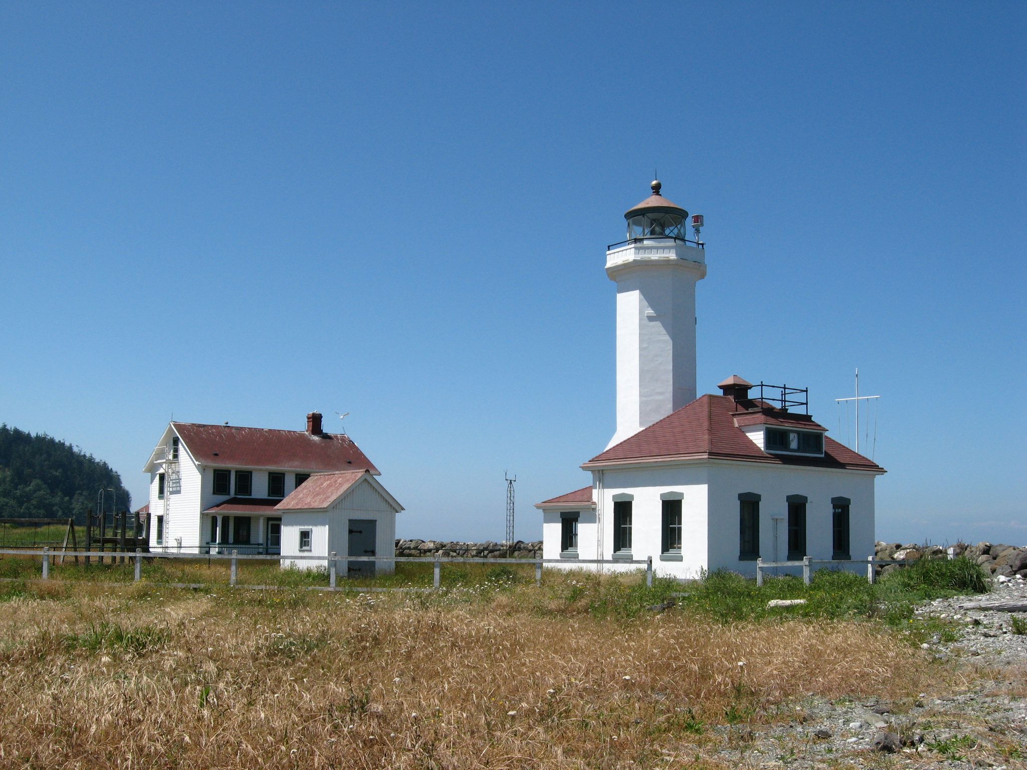 White lighthouse and buildings under a clear blue sky on a sunny day. - Point Wilson Trail