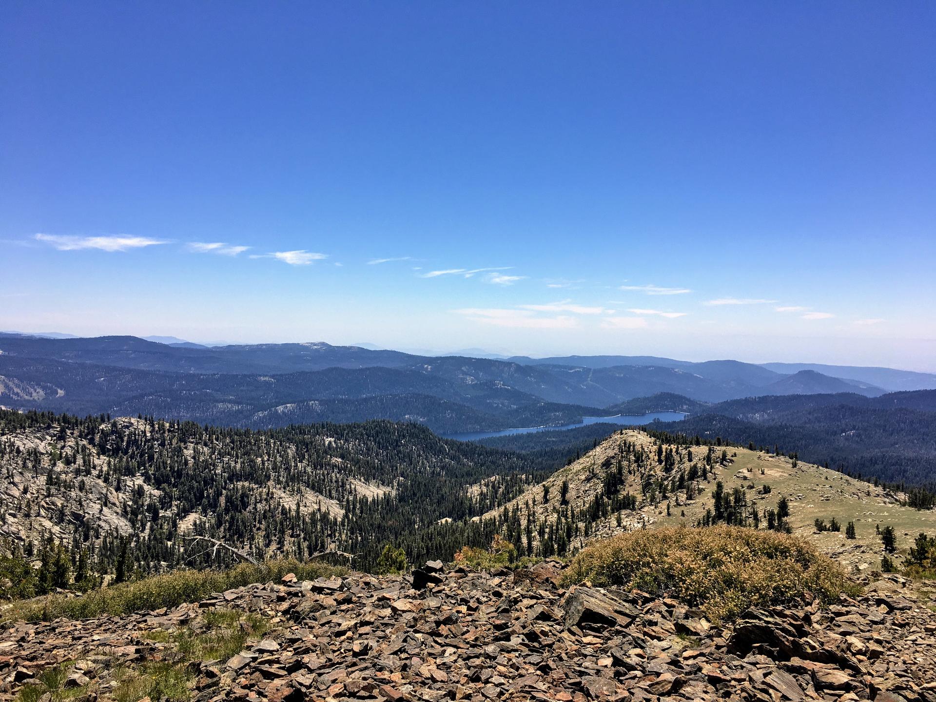 Expansive view of forested mountains and a distant lake under a clear blue sky.