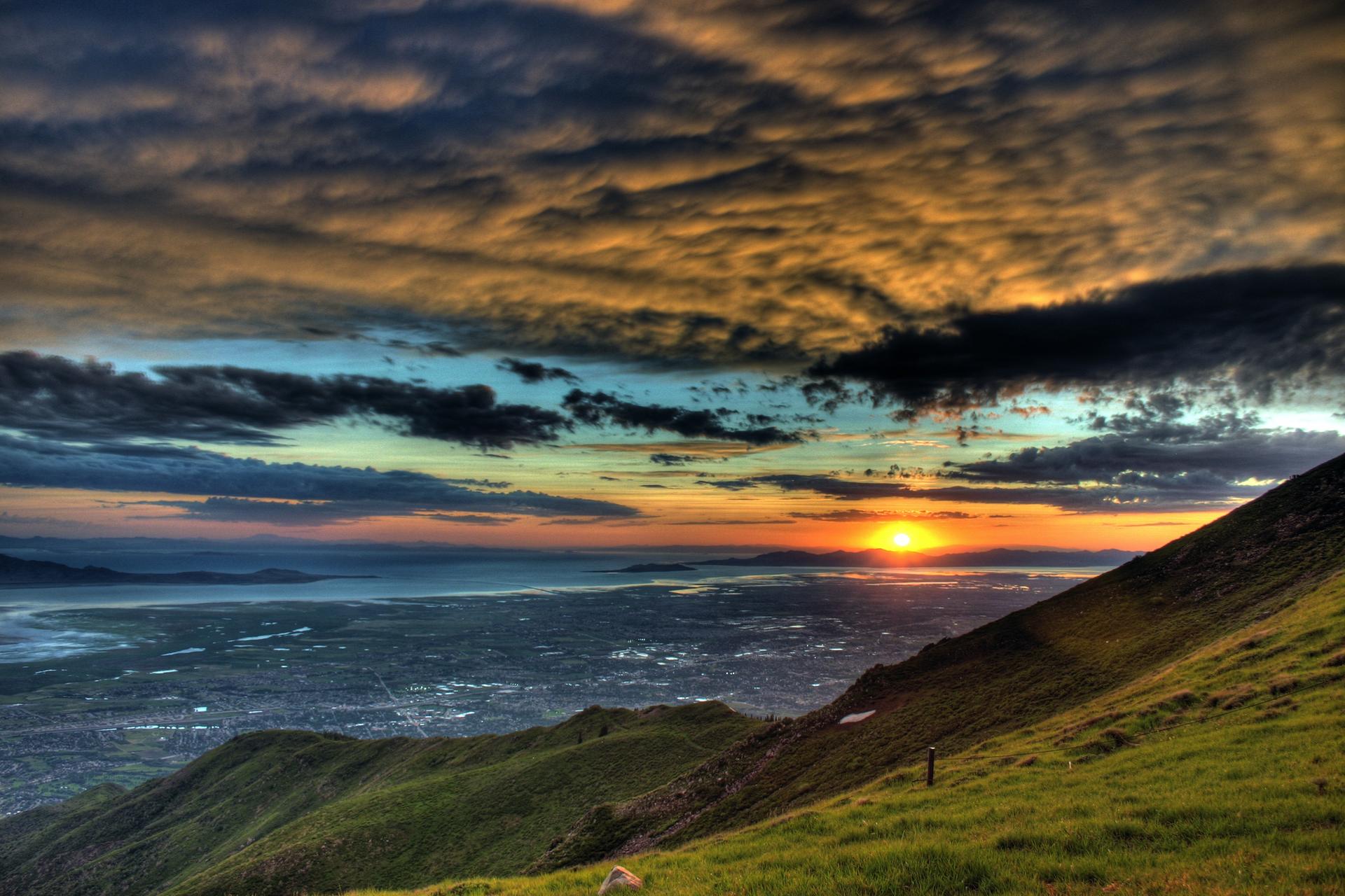 Dramatic sunset over green hills, a city, and distant water under colorful clouds.