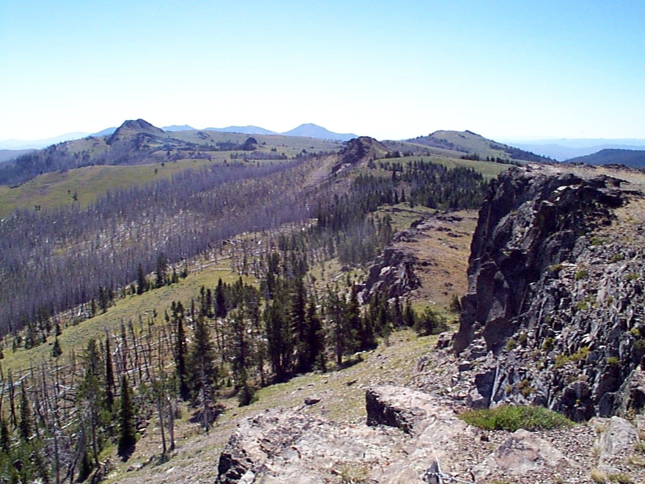 Expansive mountain landscape with rocky cliffs, green forests, and distant peaks under clear sky.