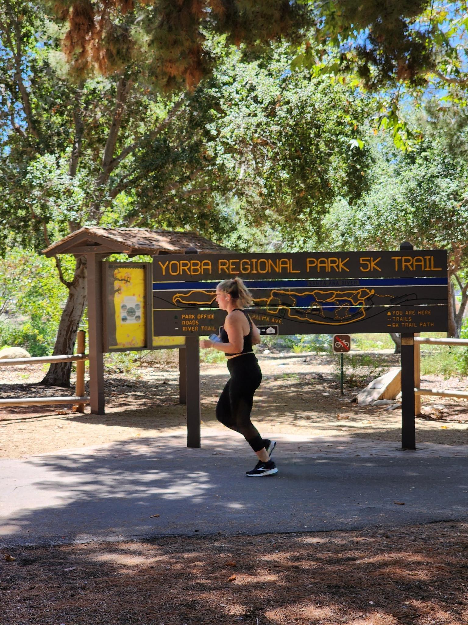 Woman running past a large trail map sign on a bright, sunny day.