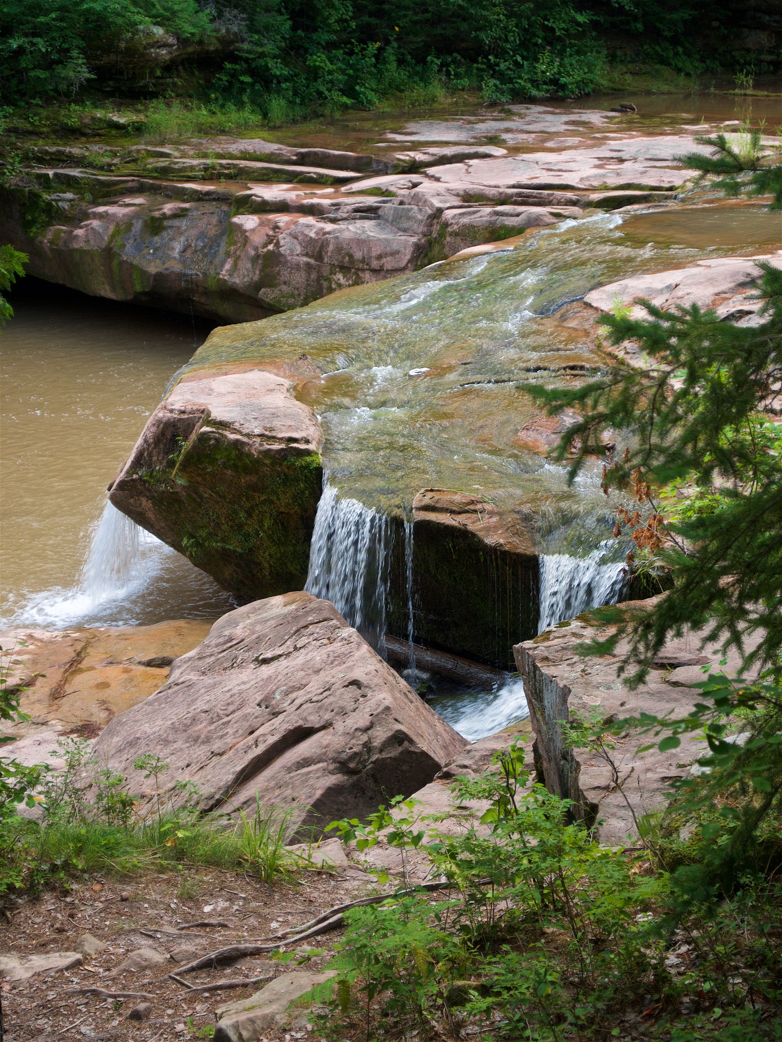 Water flows over and under large flat rocks in a riverbed with green foliage. - O Kun de Kun Falls via North Country Trail