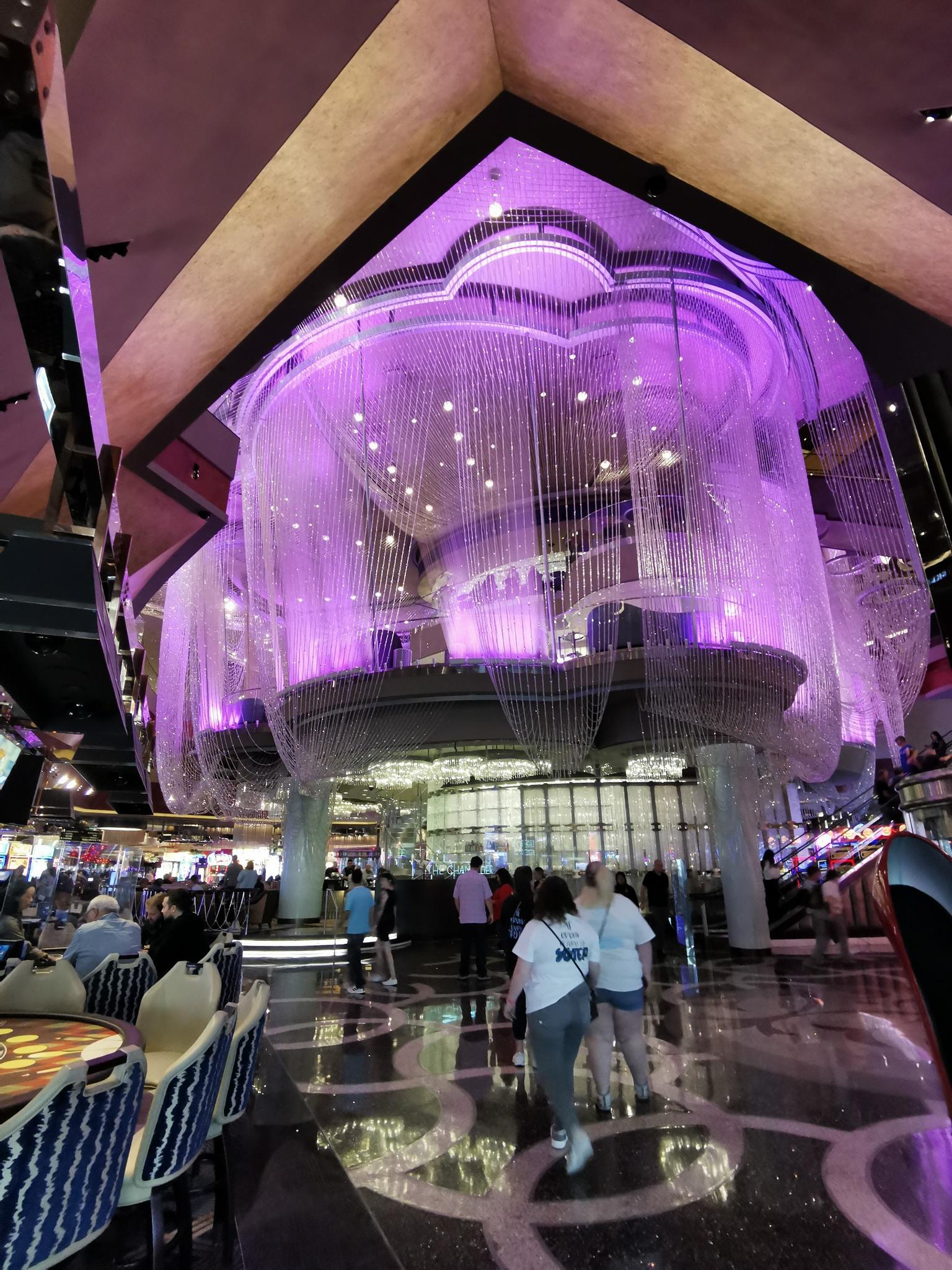 People walk under a massive, glowing purple chandelier in a grand hall.