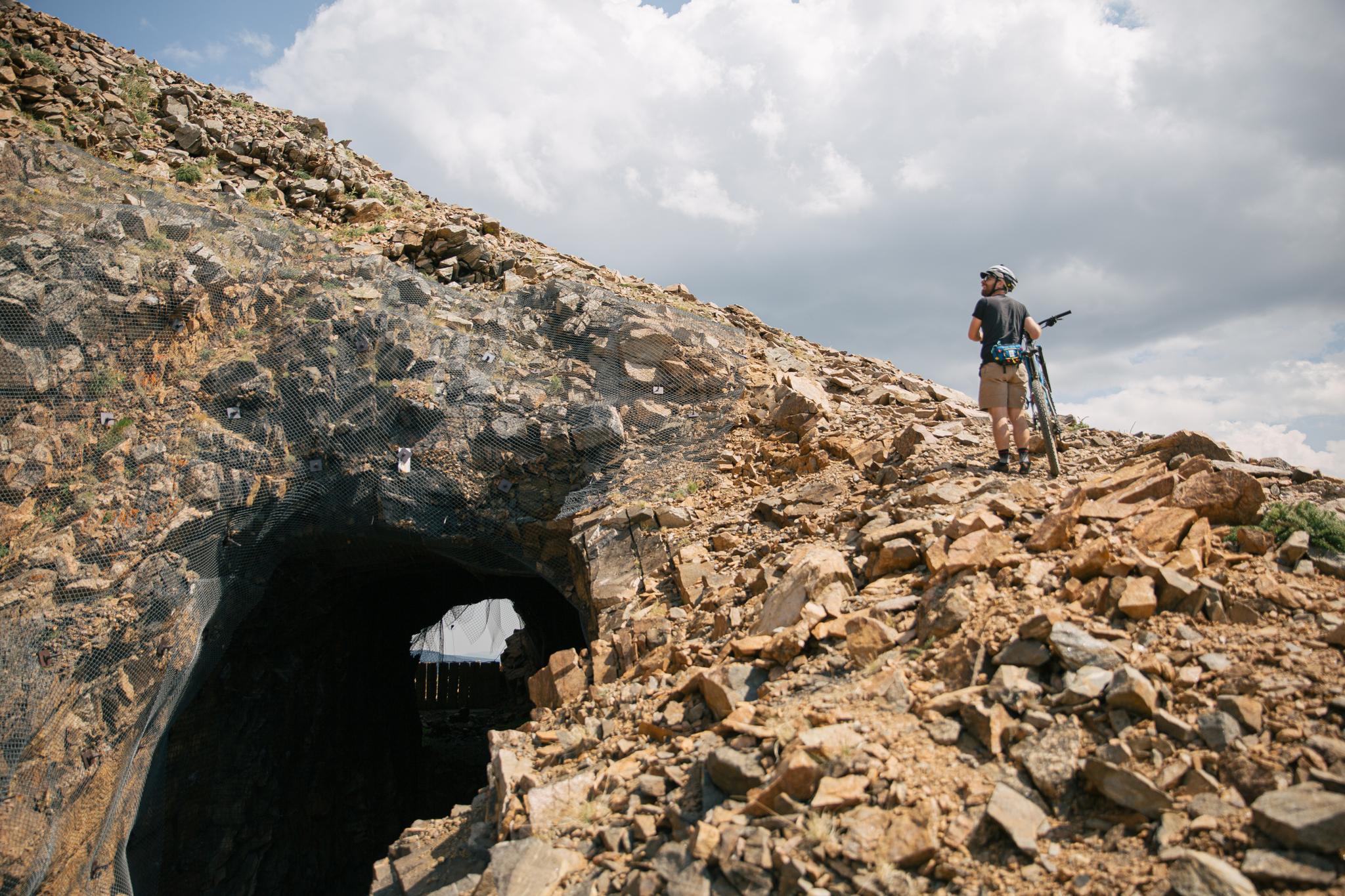 Person with bike stands on rocky slope near a dark tunnel entrance.
