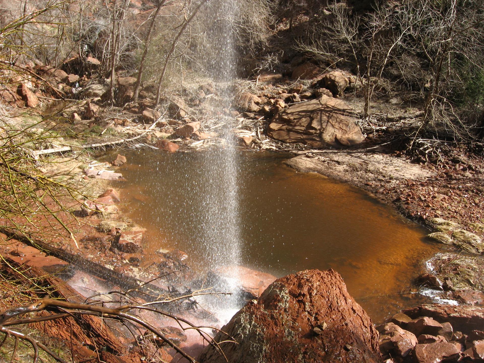 A waterfall plunges into a reddish-brown pool surrounded by rocks and sparse trees.