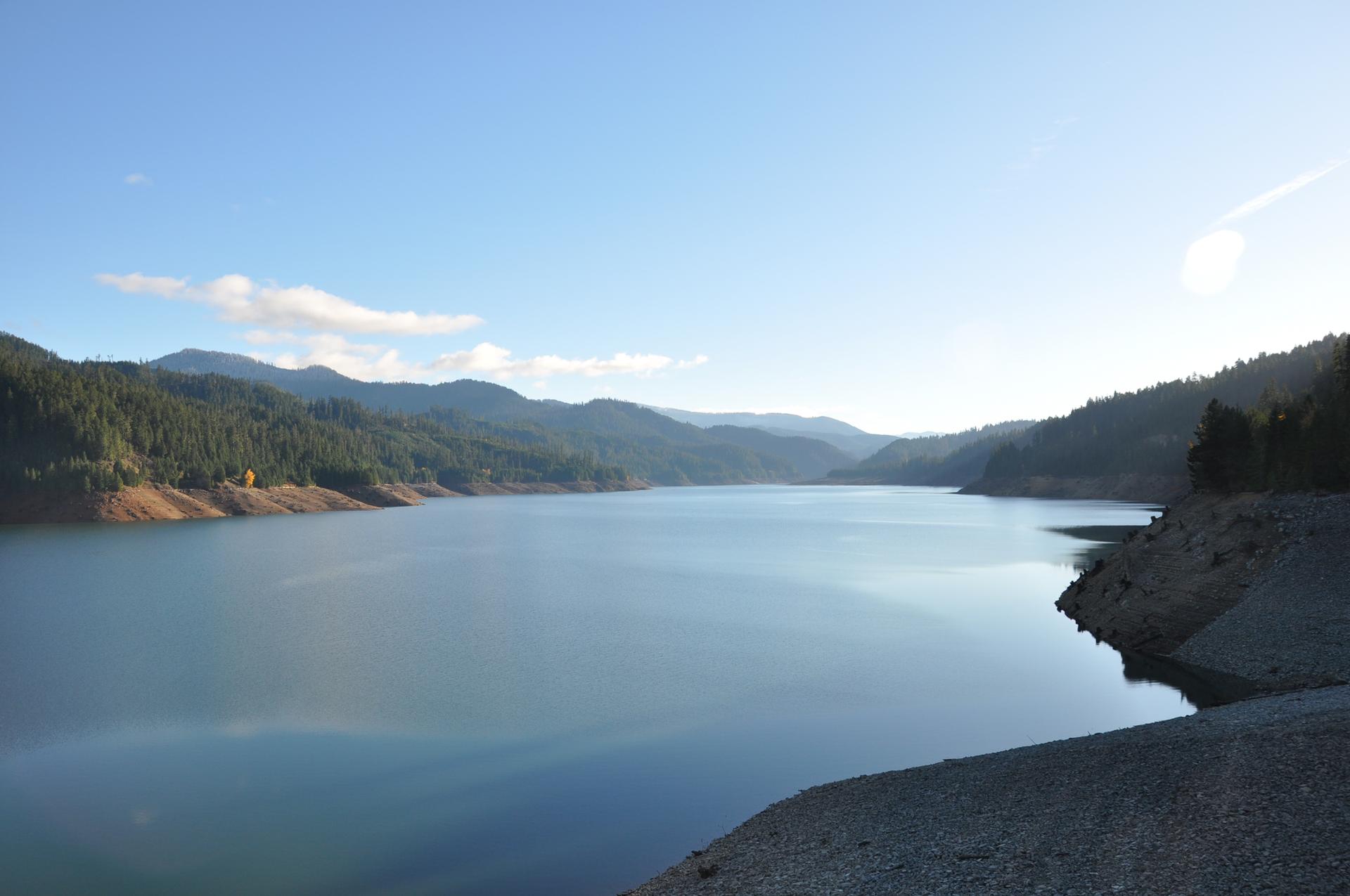 Serene lake reflecting a clear sky, surrounded by forested hills and distant mountains.