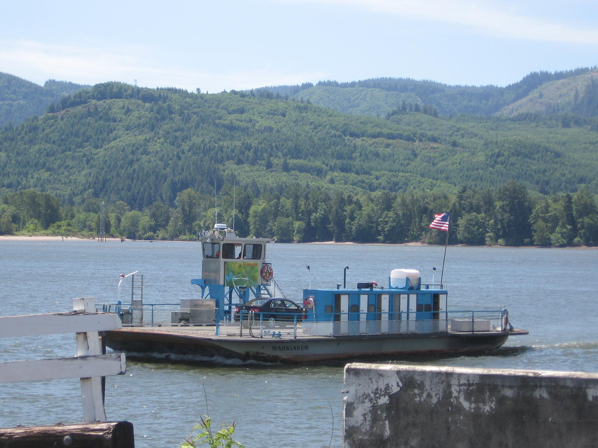 A blue and white ferry boat crosses water with forested hills behind.