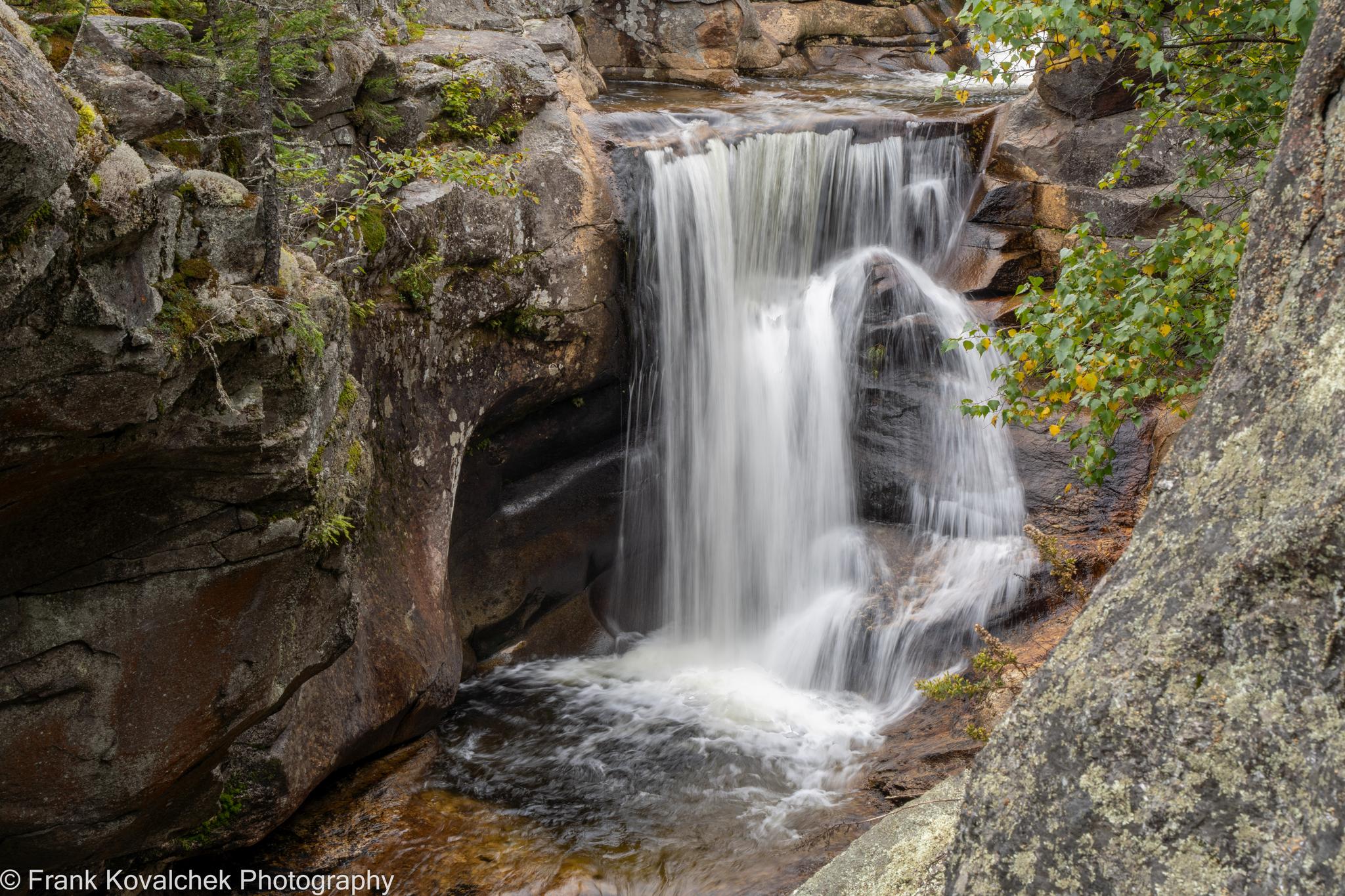 Waterfall plunges into a rocky basin with green trees under soft light.