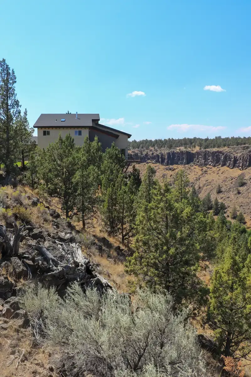 Modern house on a sunny cliff overlooking a dry, tree-filled canyon.