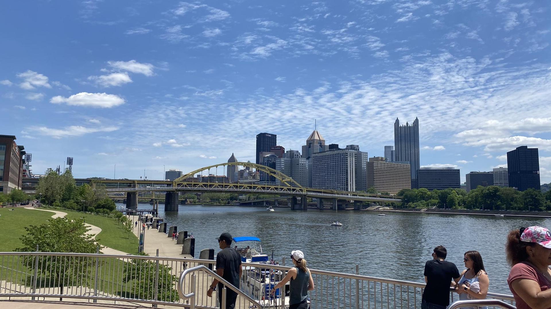 People on a sunny riverfront overlook with a city skyline and bridge.