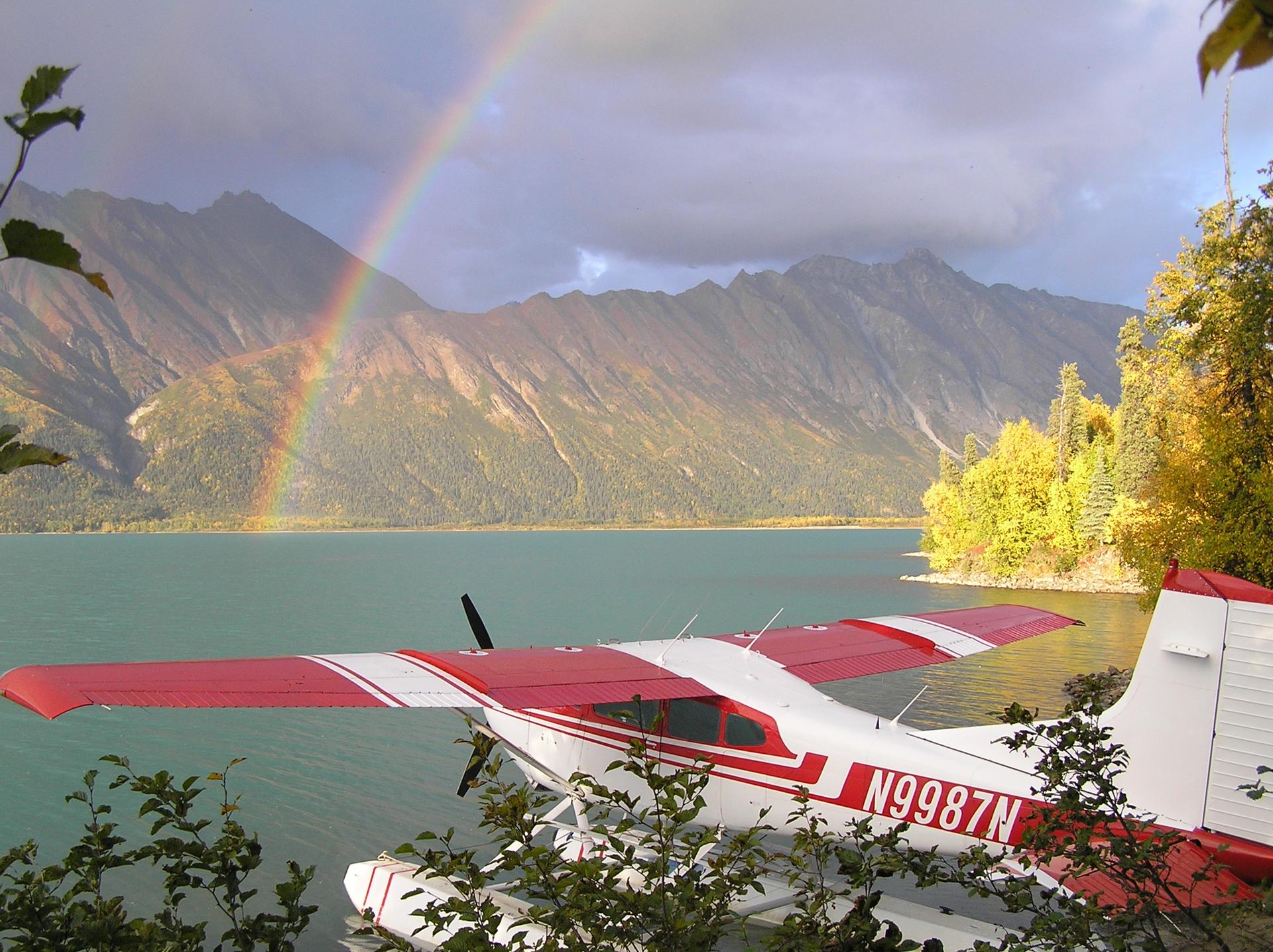 A red and white floatplane sits on a lake with a rainbow over mountains.