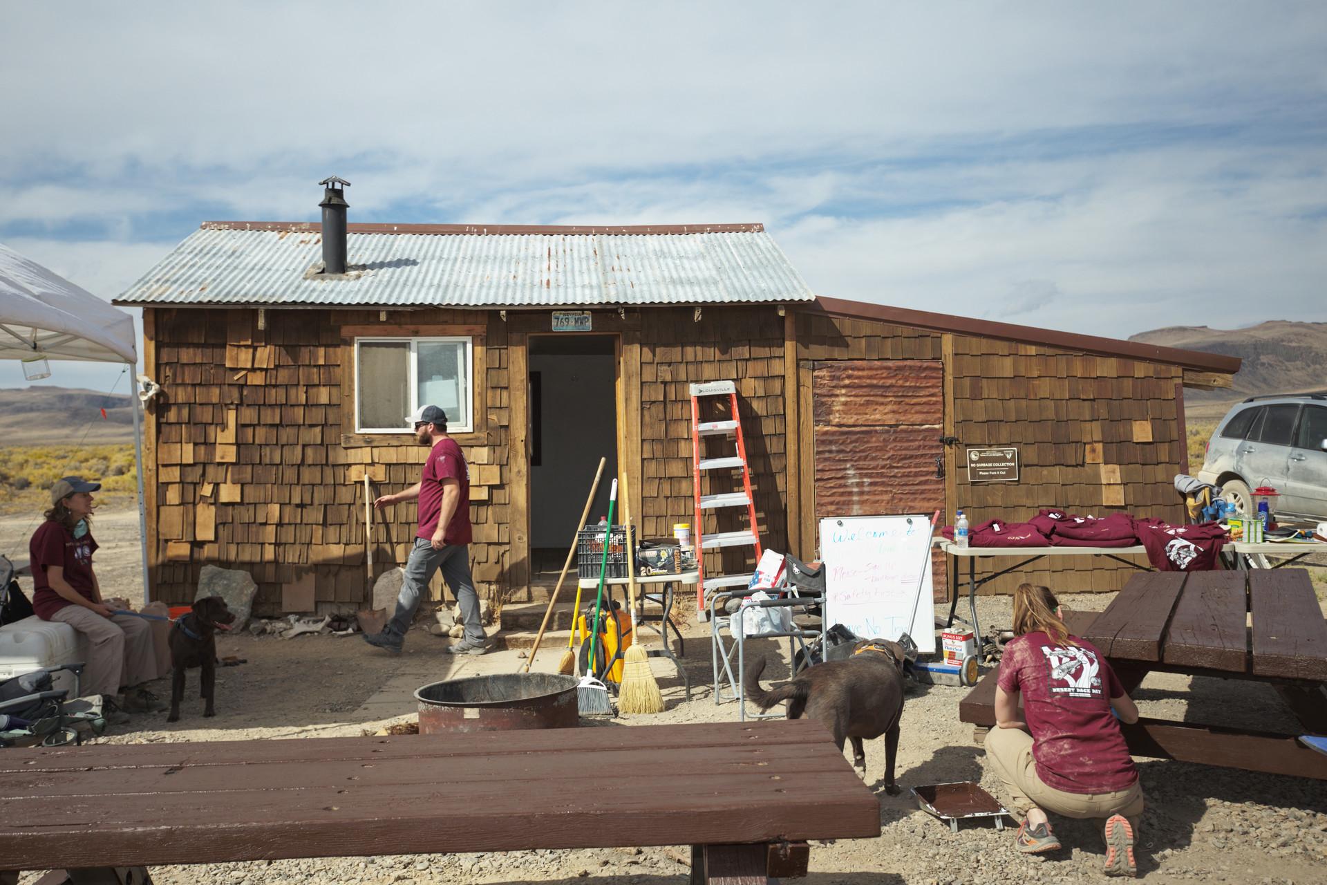 People and dogs outside a rustic wooden cabin in a sunny, arid landscape.
