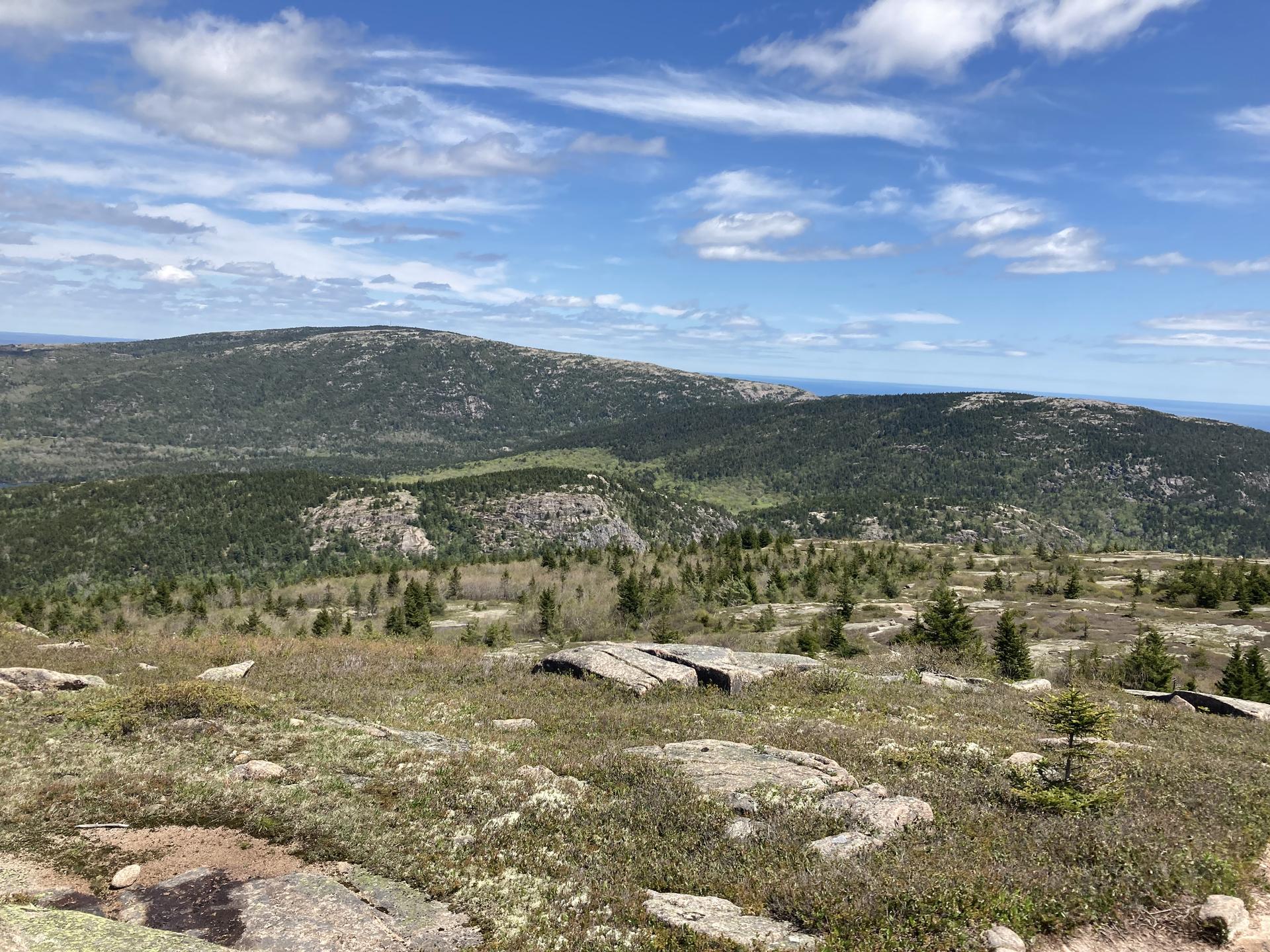 Sunny day with clouds, vast rocky and grassy landscape with distant forested mountains.