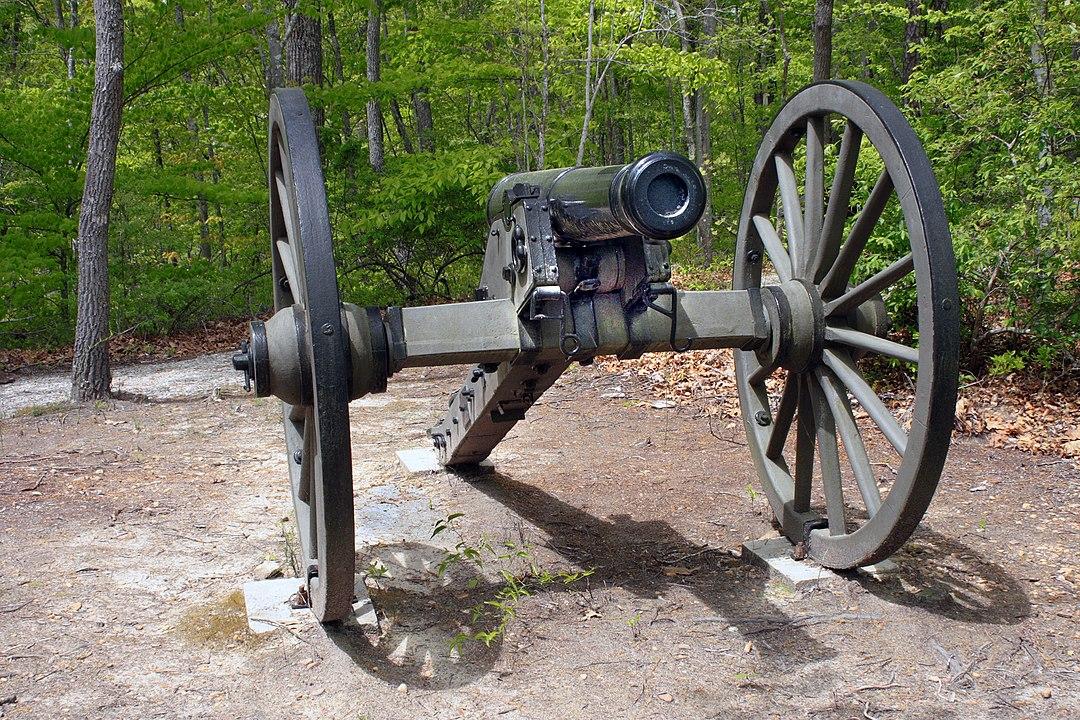 An old cannon with large wooden wheels sits in a sunny forest.