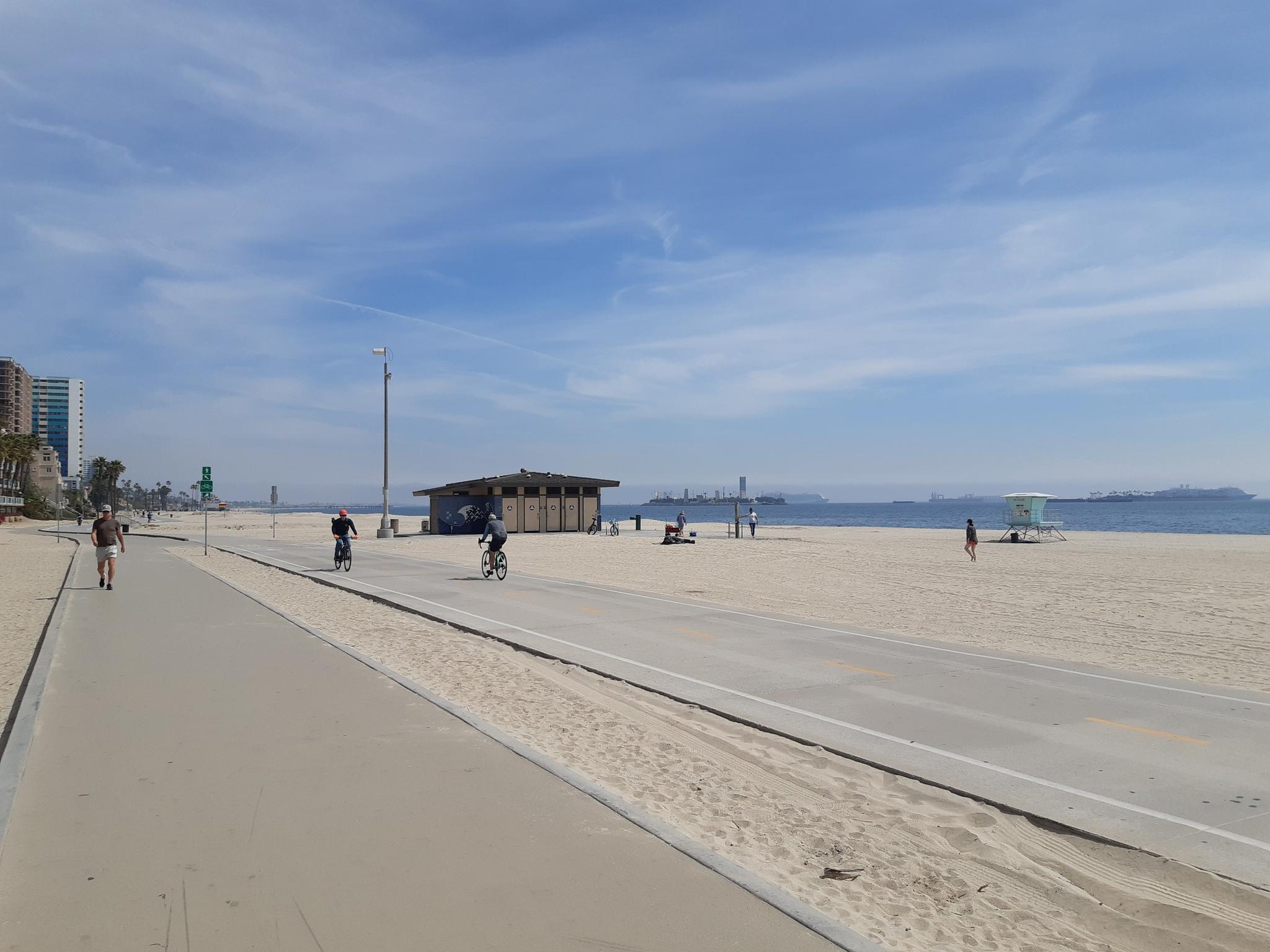Sunny beach with a paved path, people walking and cycling, and distant ocean.
