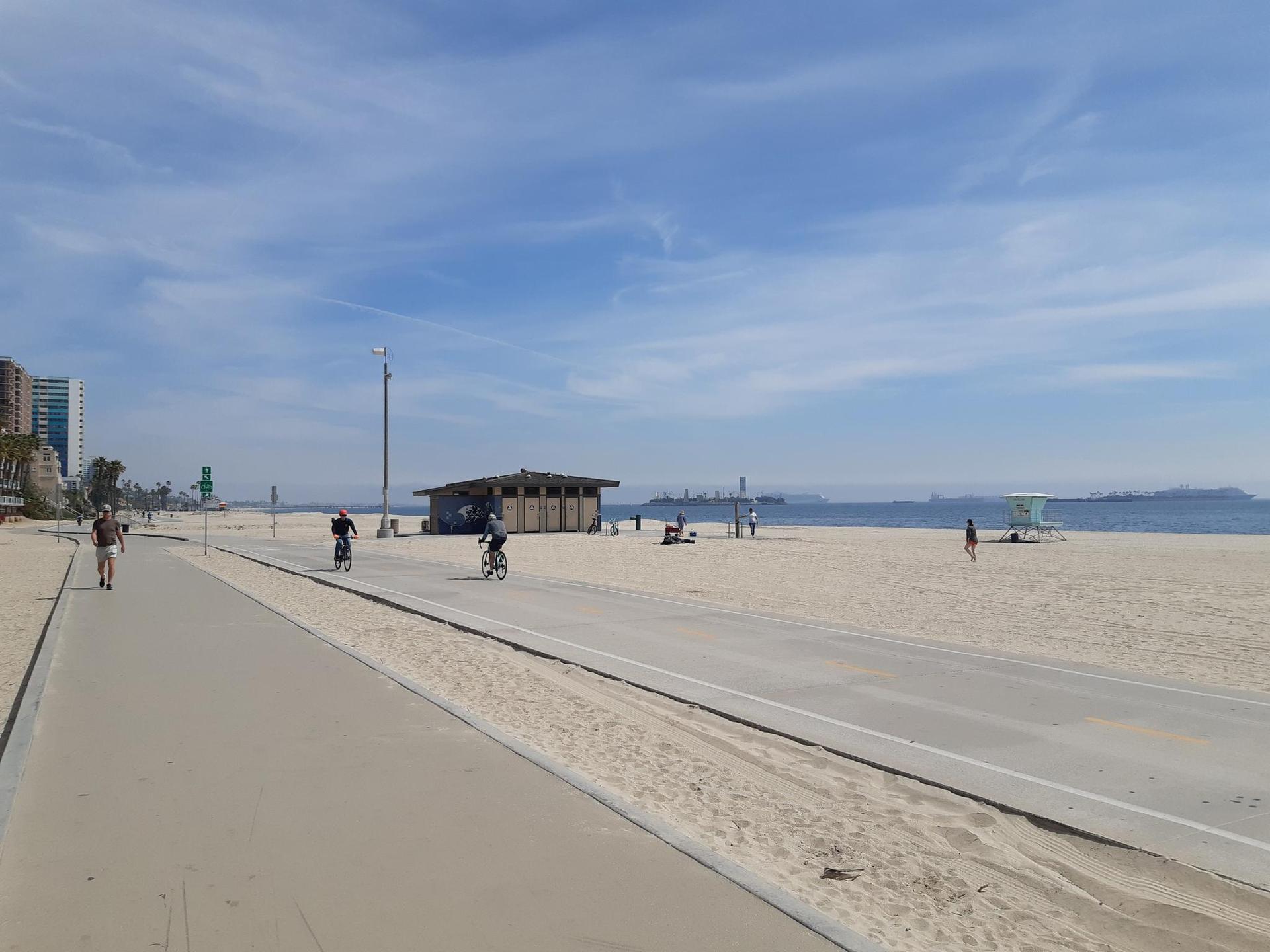 Sunny beach with a paved path, people walking and cycling, and distant ocean.