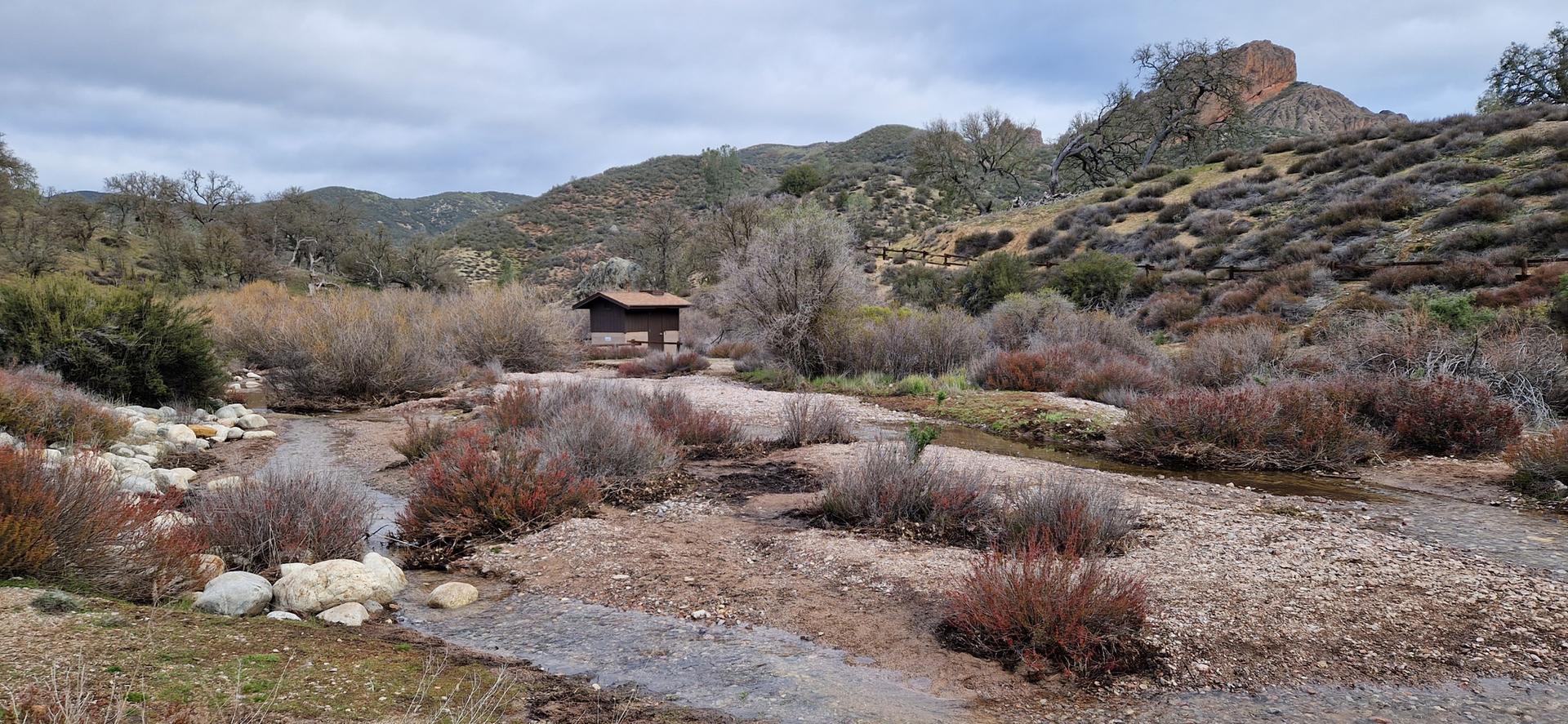 A small stream flows through a scrubby valley with a wooden shelter and hills.
