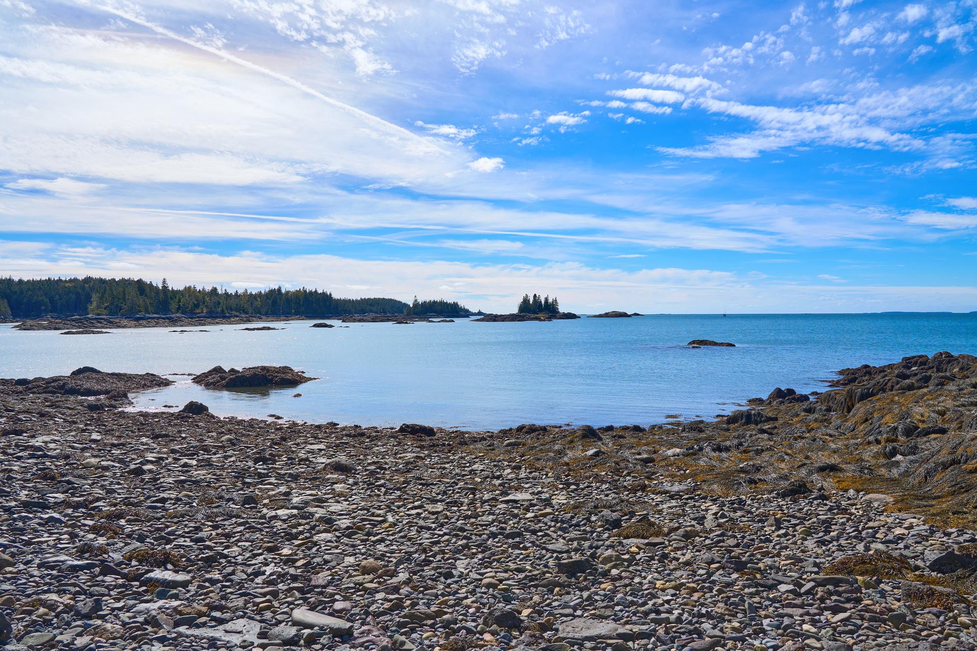 Rocky beach leading to calm blue ocean with distant forested islands under a bright sky.