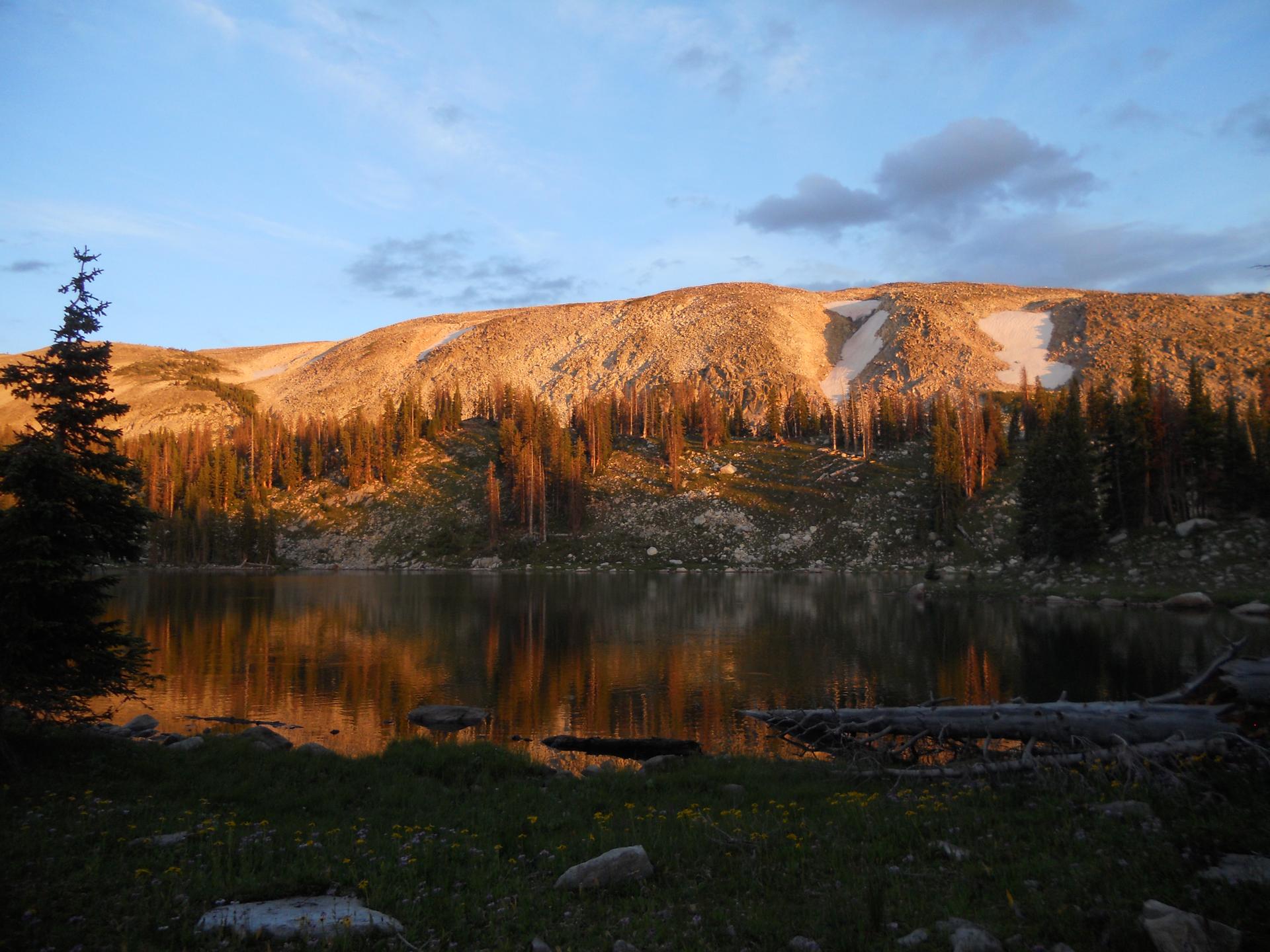 Golden light illuminates a mountain lake, pine trees, and rocky slopes with snow.