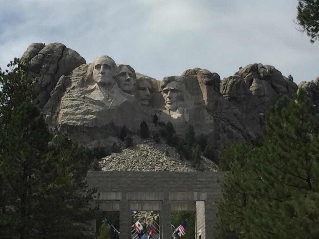 Four large faces carved into a rocky mountain, framed by pine trees.