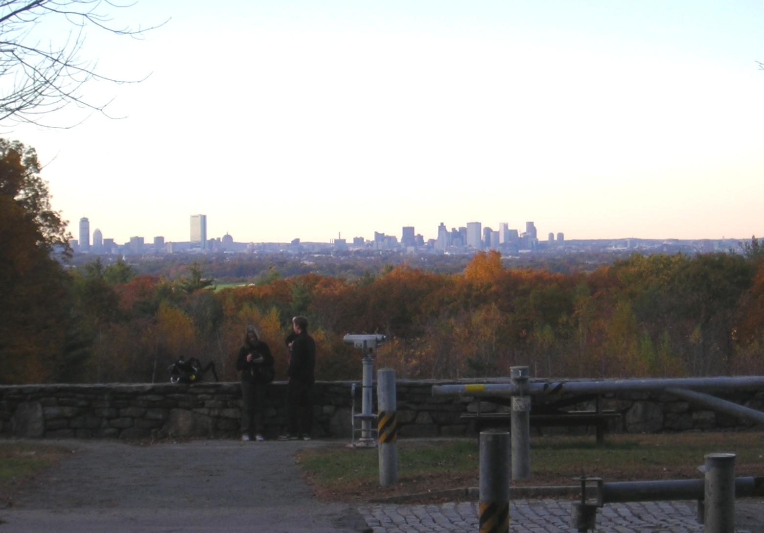 Two people at an overlook with autumn trees and a distant city skyline.
