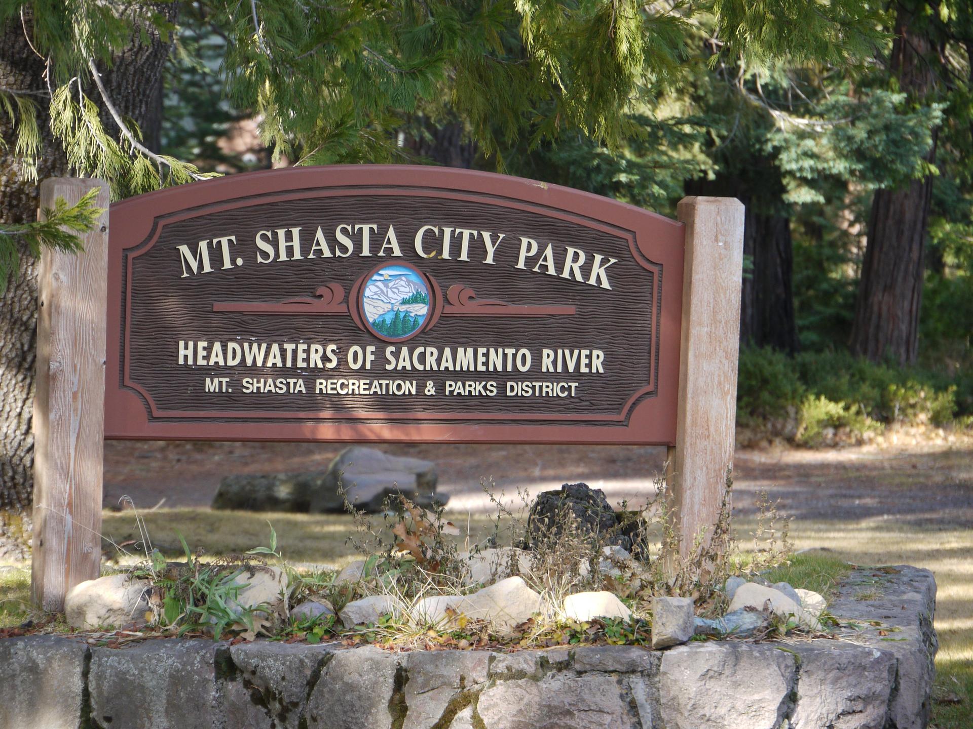 A wooden park sign on a stone base, surrounded by trees in bright sunlight.