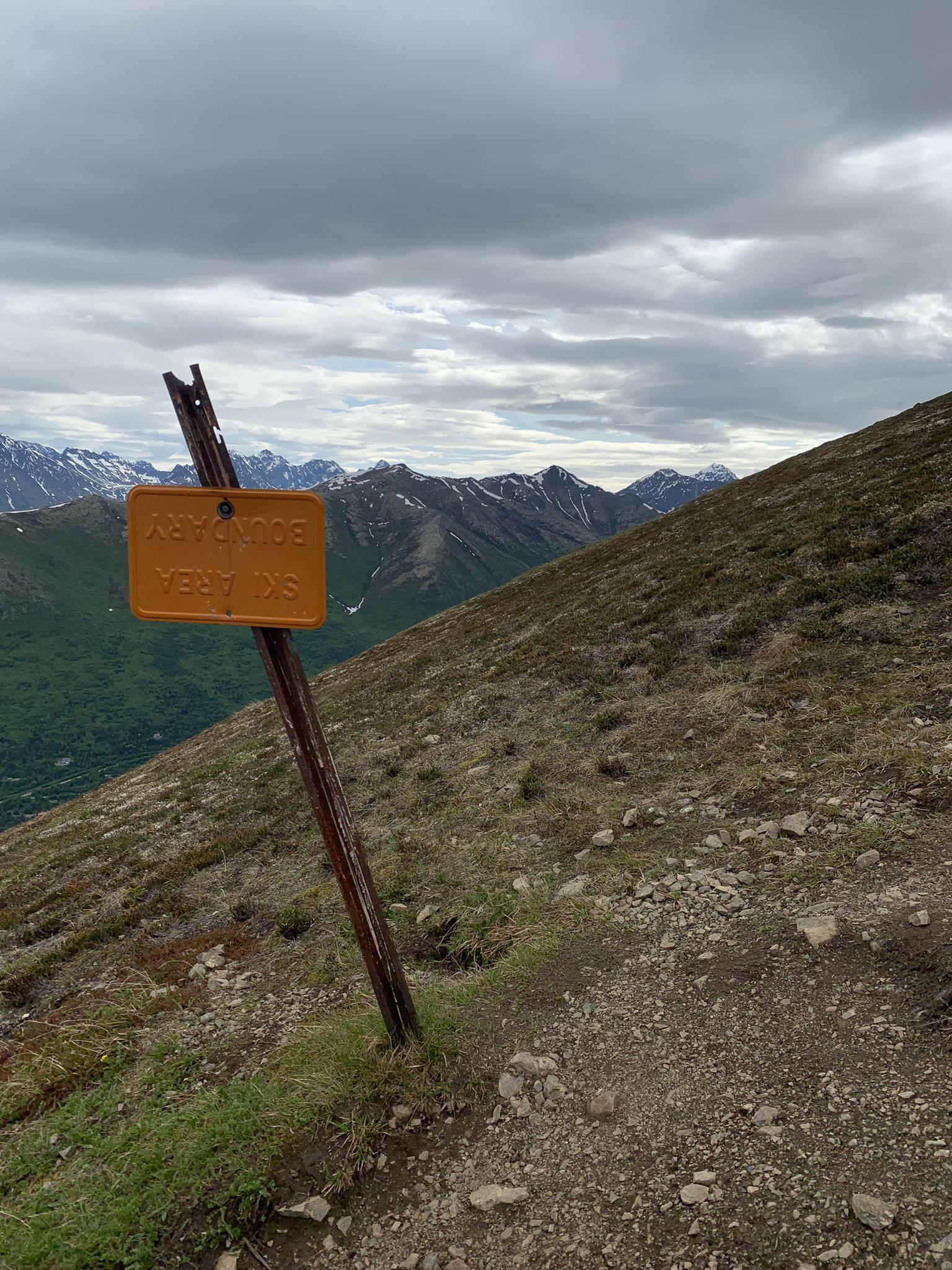 An orange signpost on a steep, rocky mountain trail under cloudy skies.