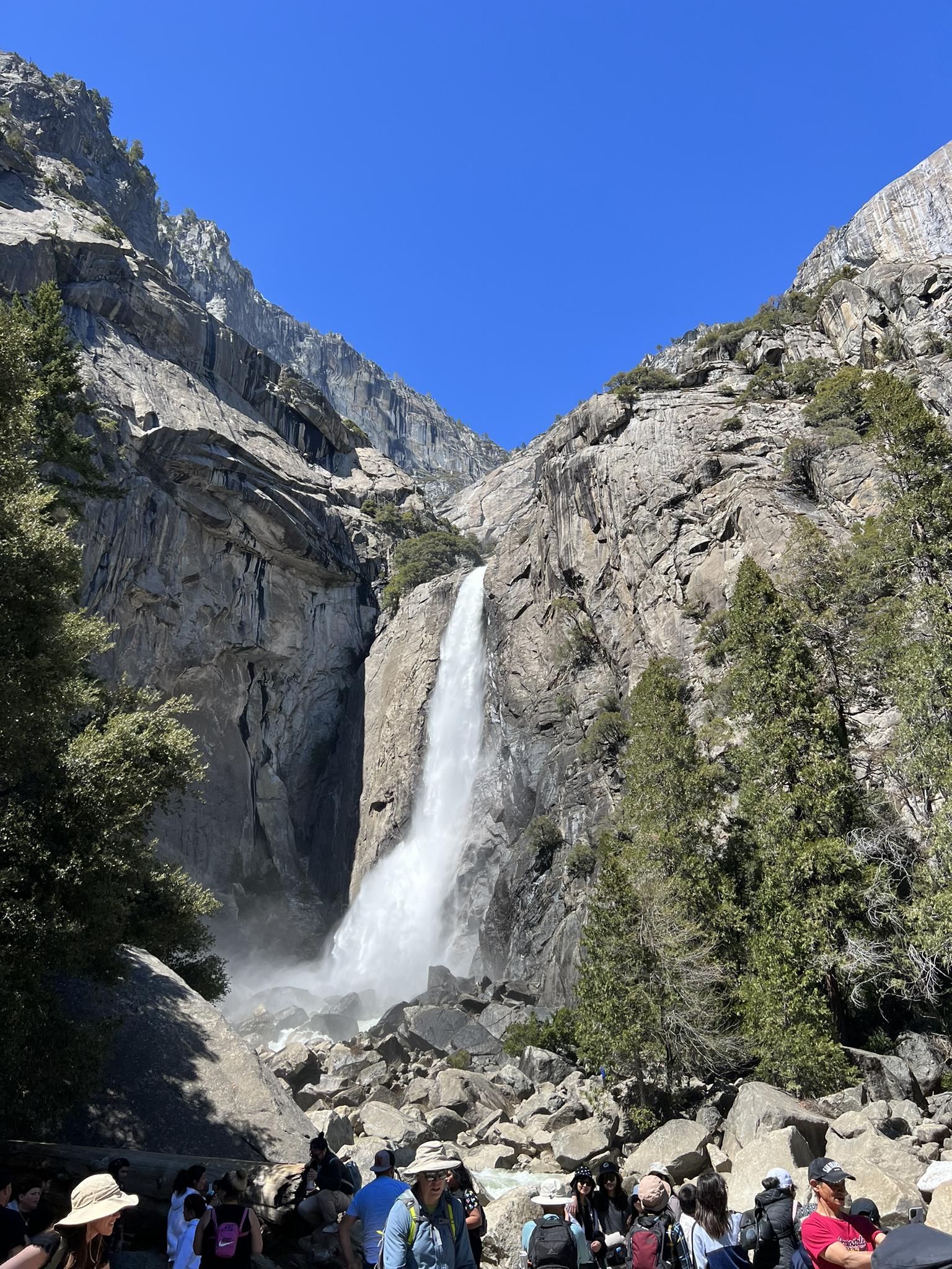 Bright daylight illuminates a powerful waterfall cascading down rocky cliffs, with many people below.