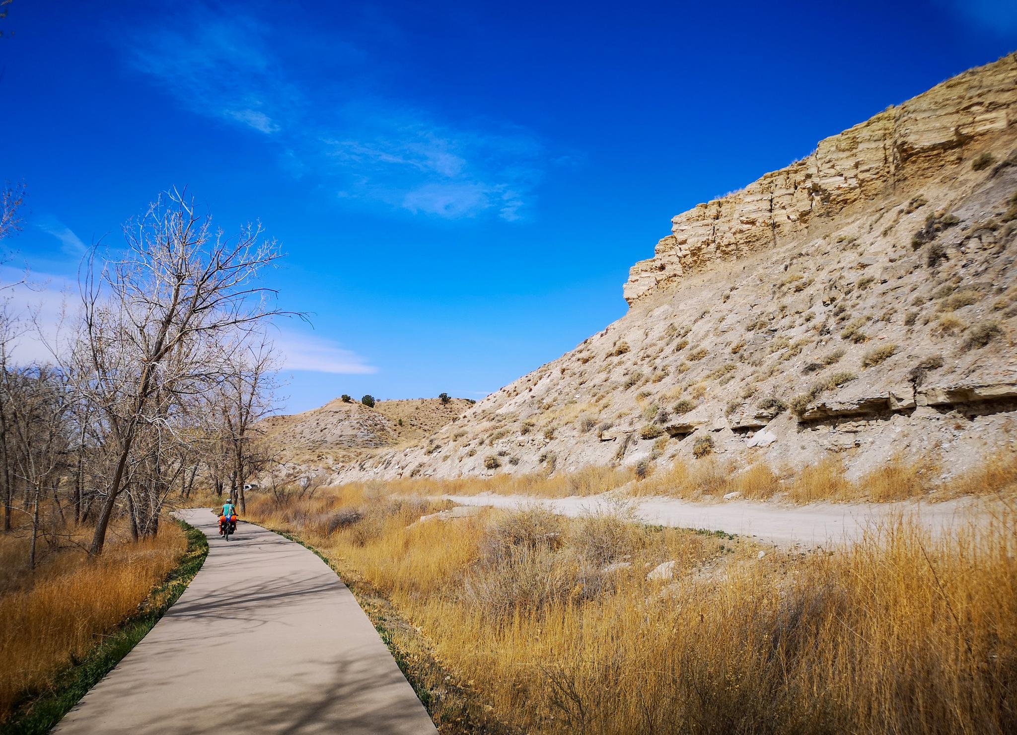 A person cycles on a paved path beside a rocky, dry hillside under blue sky.
