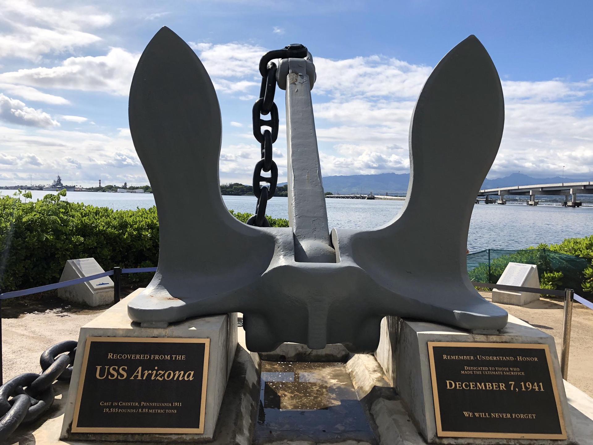 A large grey anchor with chain, plaques, water, and distant bridge under cloudy sky.