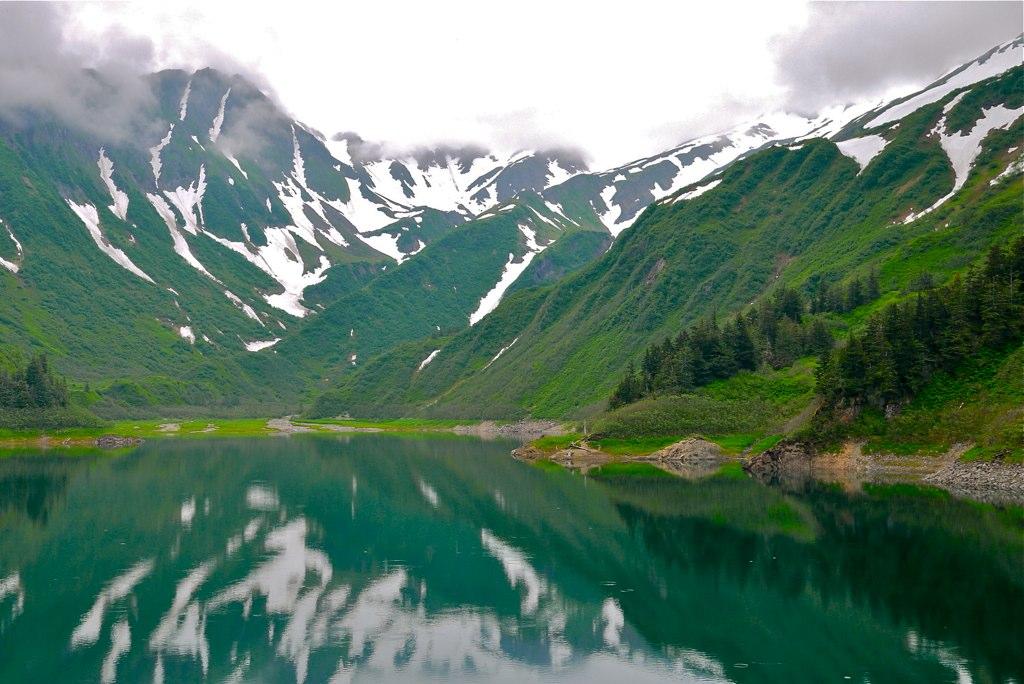Calm green lake reflects snow-capped mountains under a cloudy sky.