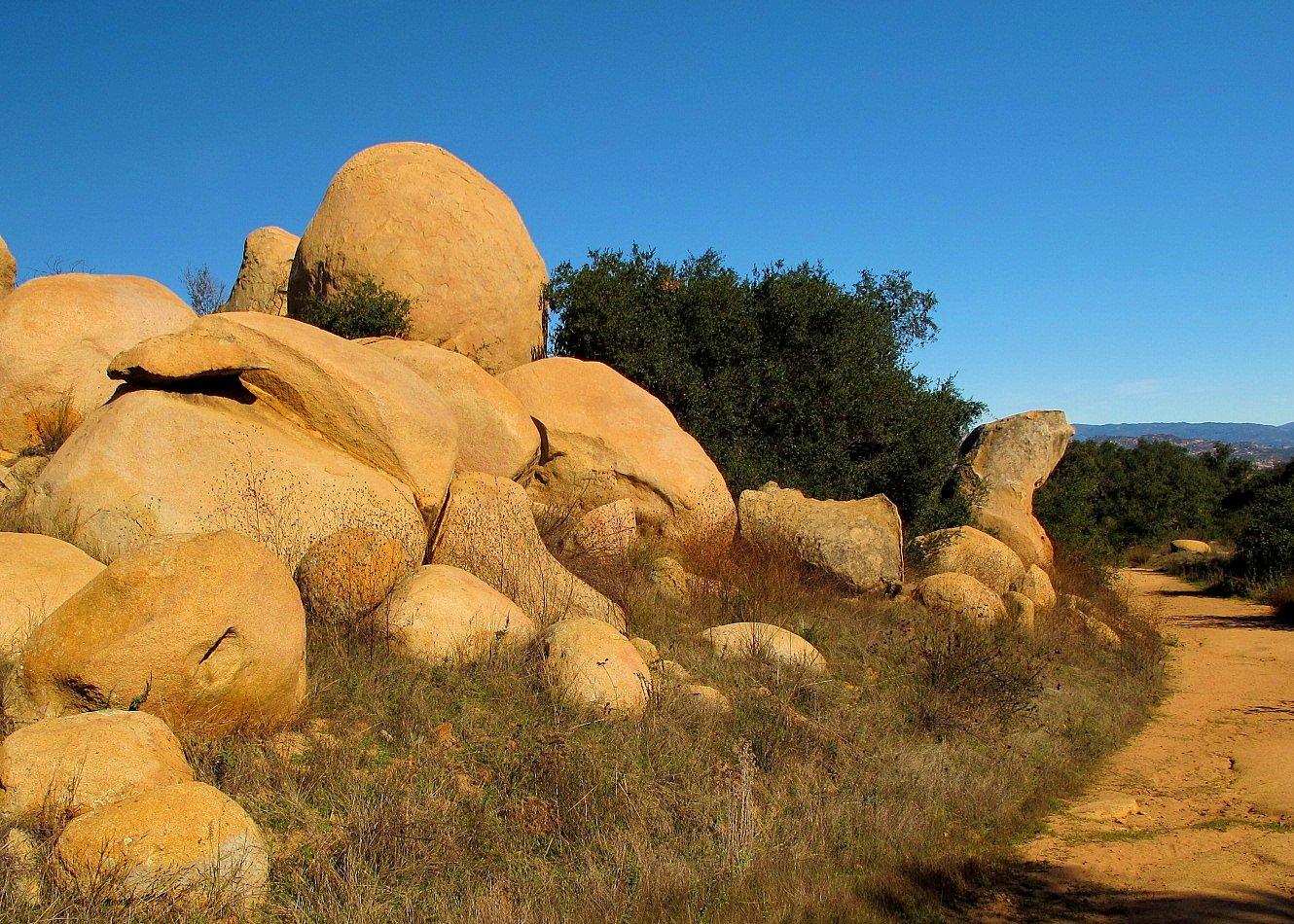 Large, sunlit boulders and green trees line a dirt path under a clear blue sky.