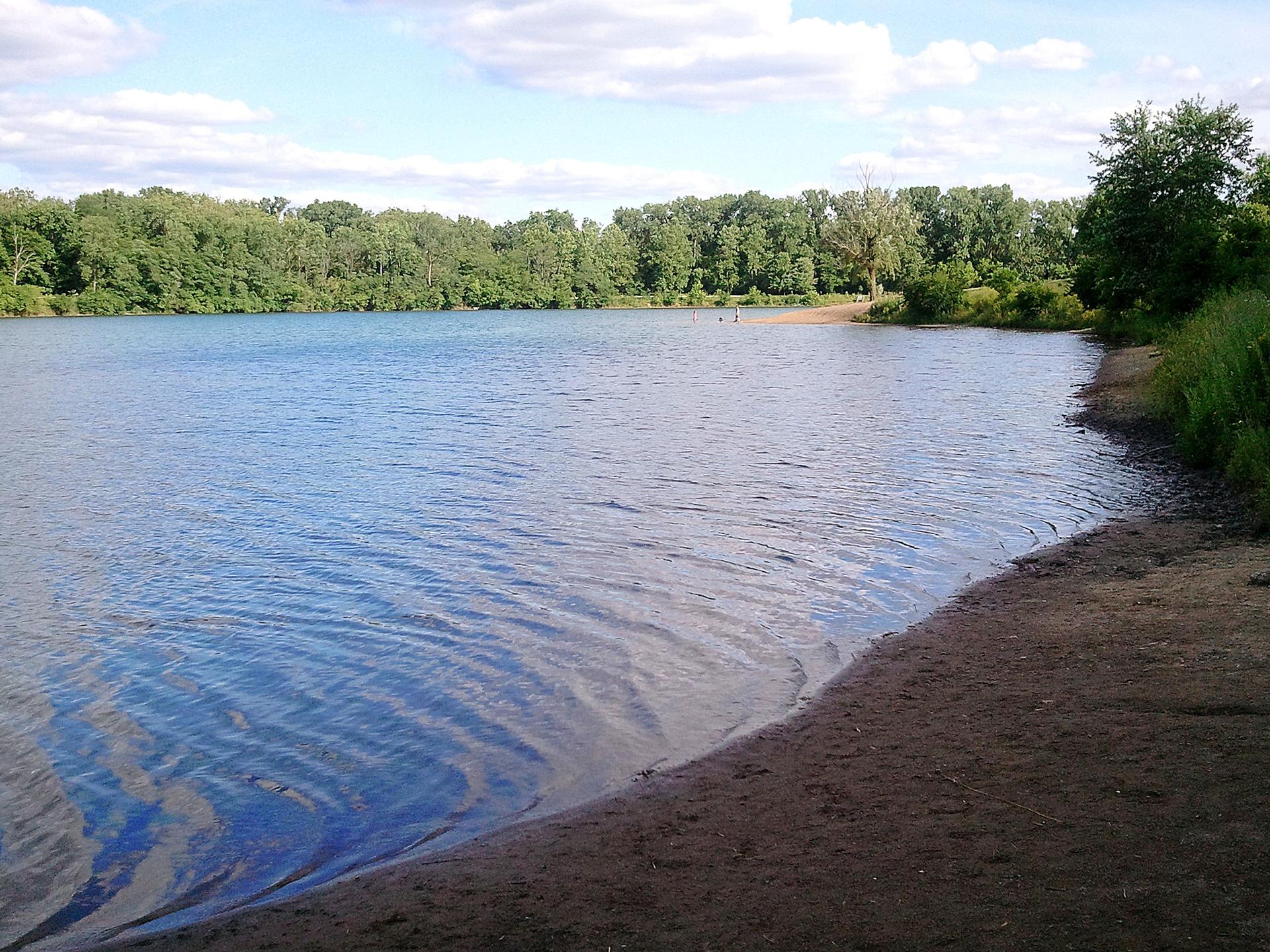 Bright lake with sandy shore, green trees, and distant people.