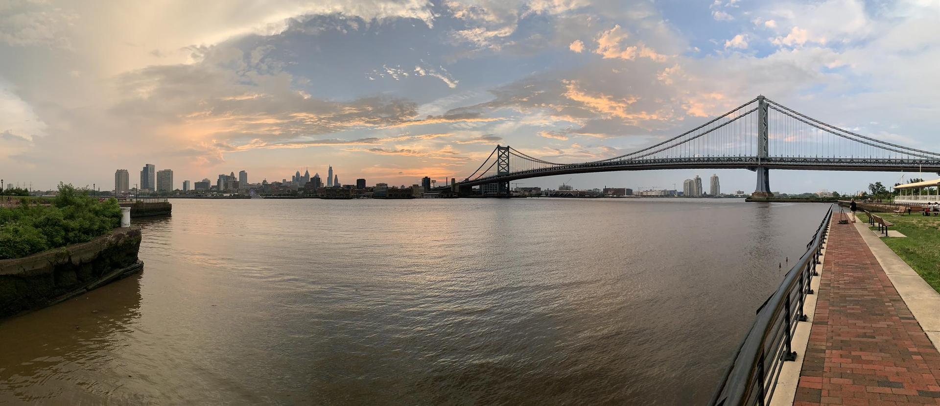 Panoramic view of a suspension bridge over a river at sunset, with a distant city skyline.