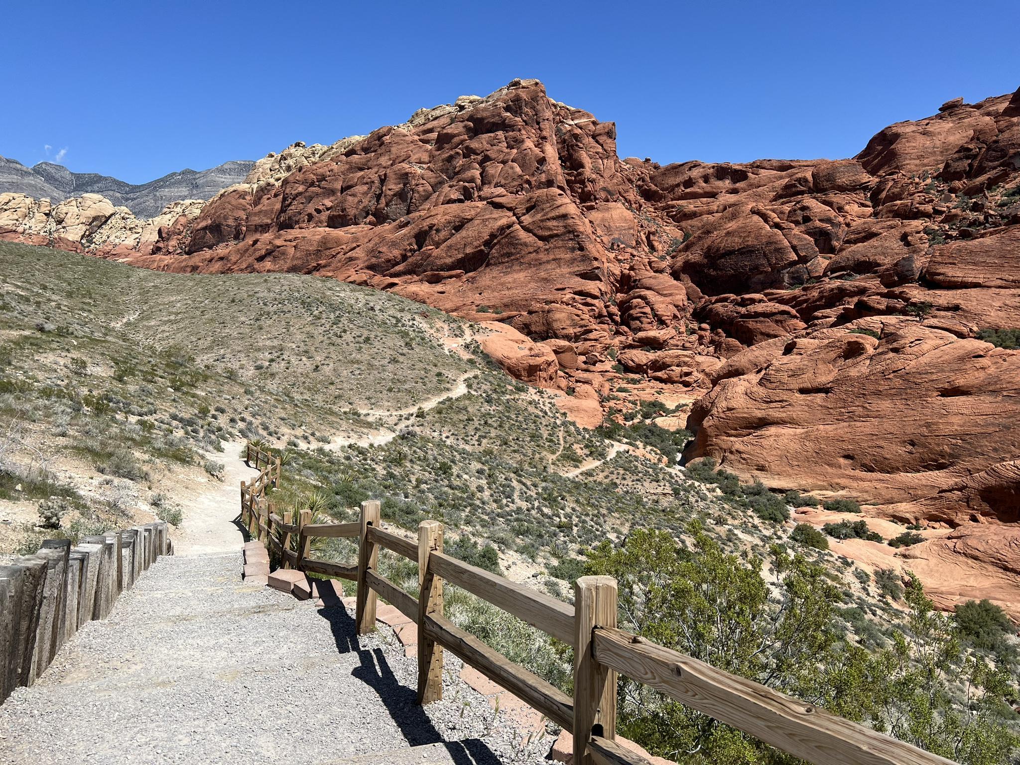 A gravel path with a wooden fence leading towards red rock mountains on a sunny day.