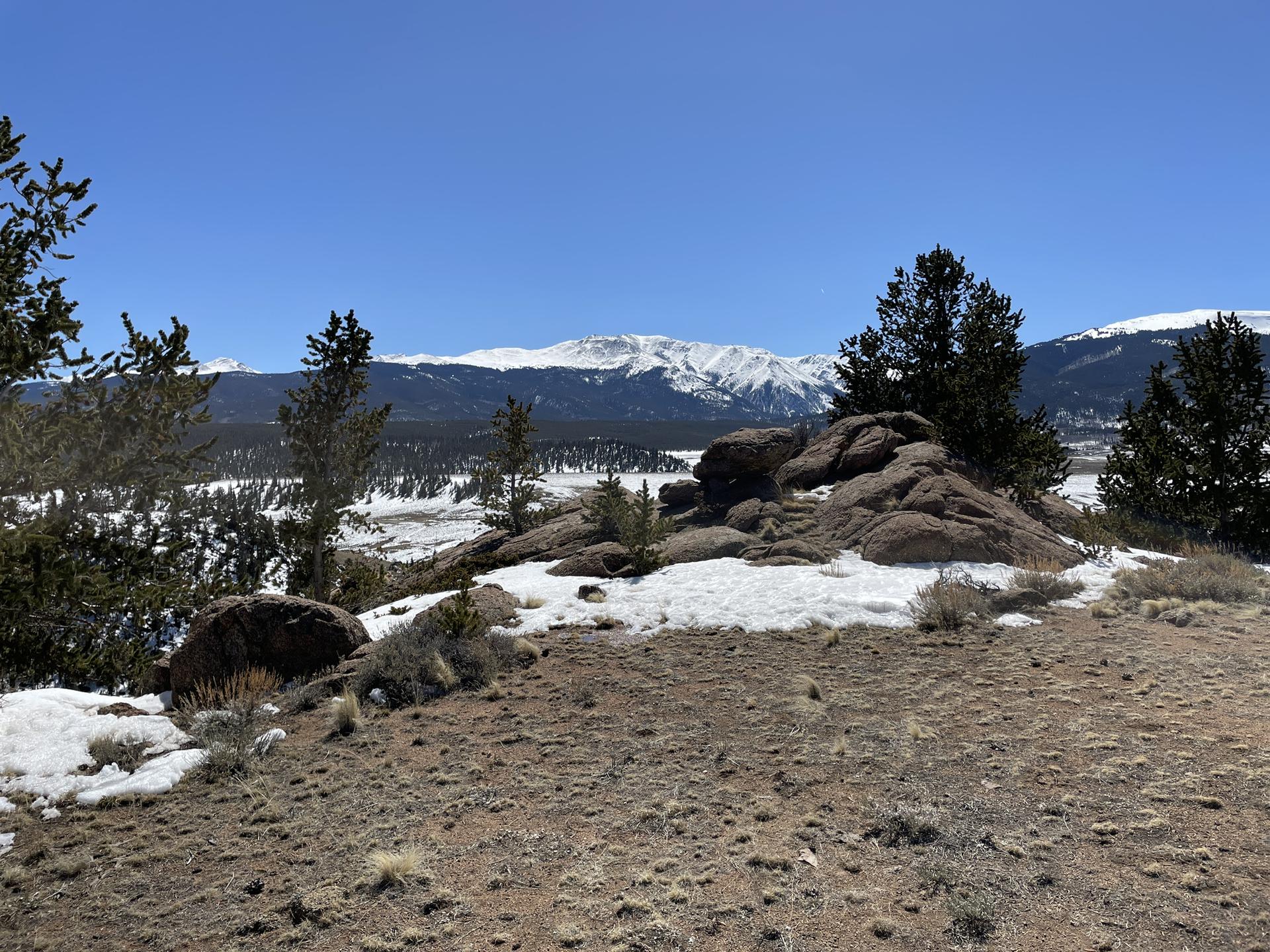 Rocky terrain and snow patches overlook a vast valley with snowy mountains.