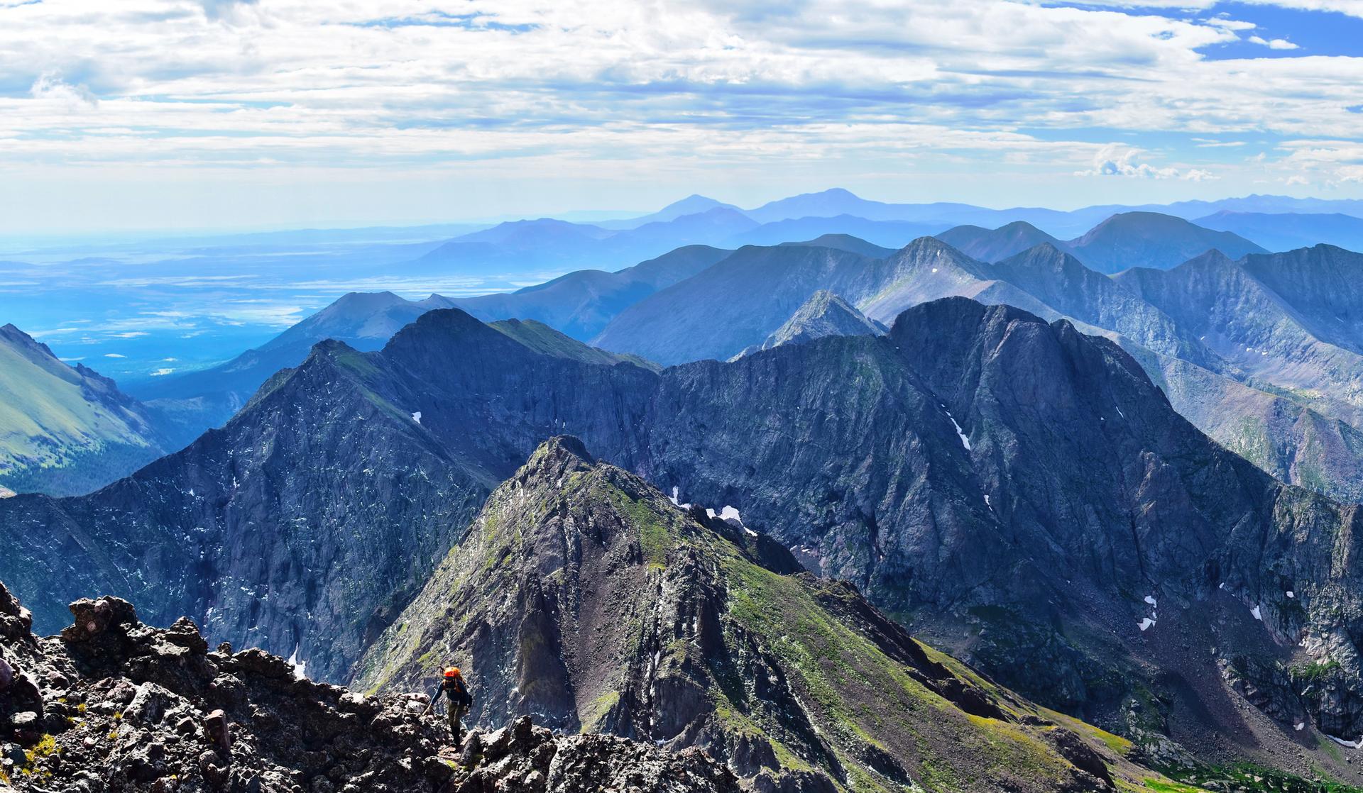 A person hikes a rocky mountain ridge under a partly cloudy sky.