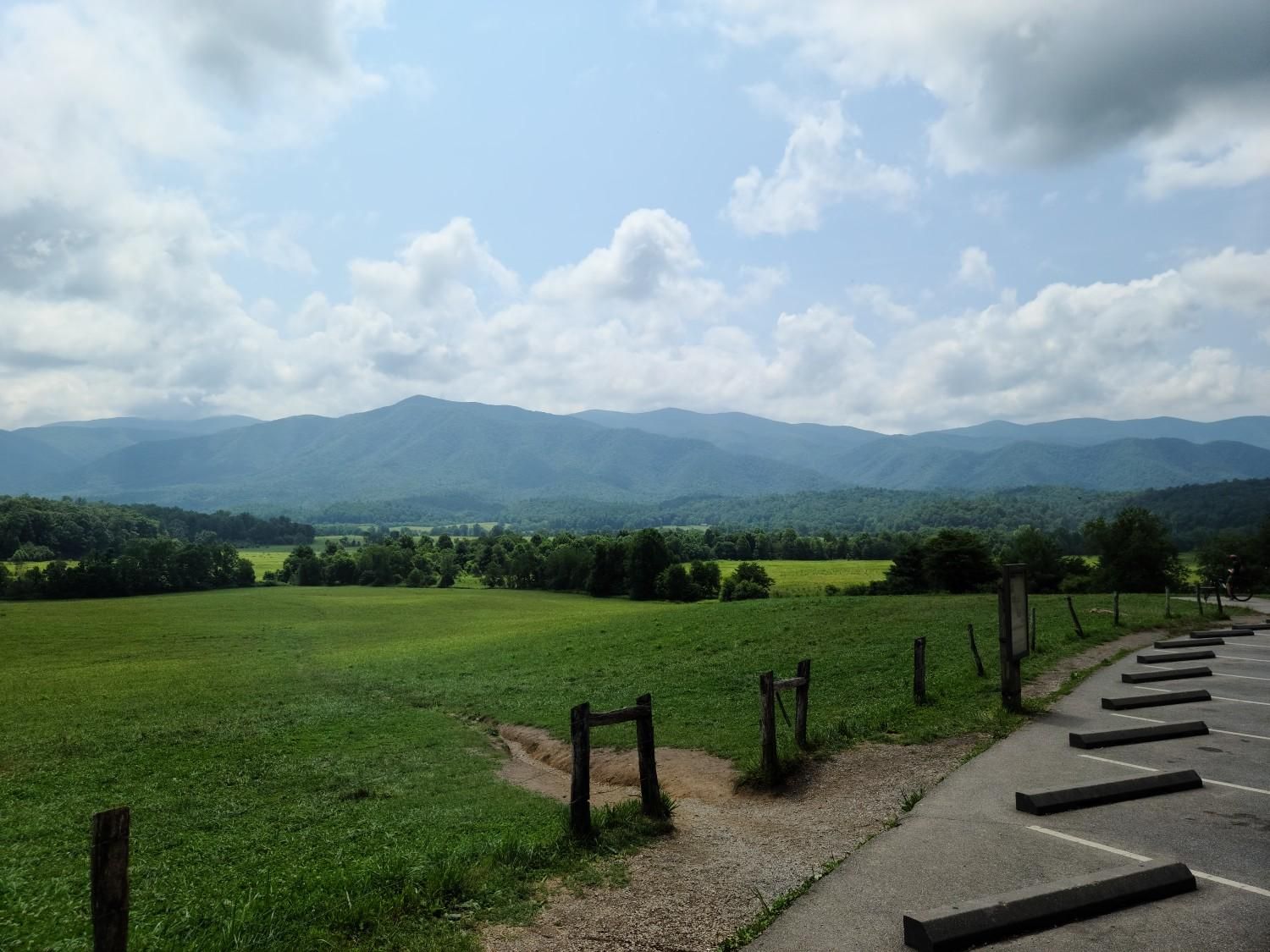 Expansive green field with distant mountains under a partly cloudy sky, near a parking lot. - Cades Cove Loop Road – Beautiful view of the Valley loop from Rich Mountain Gap