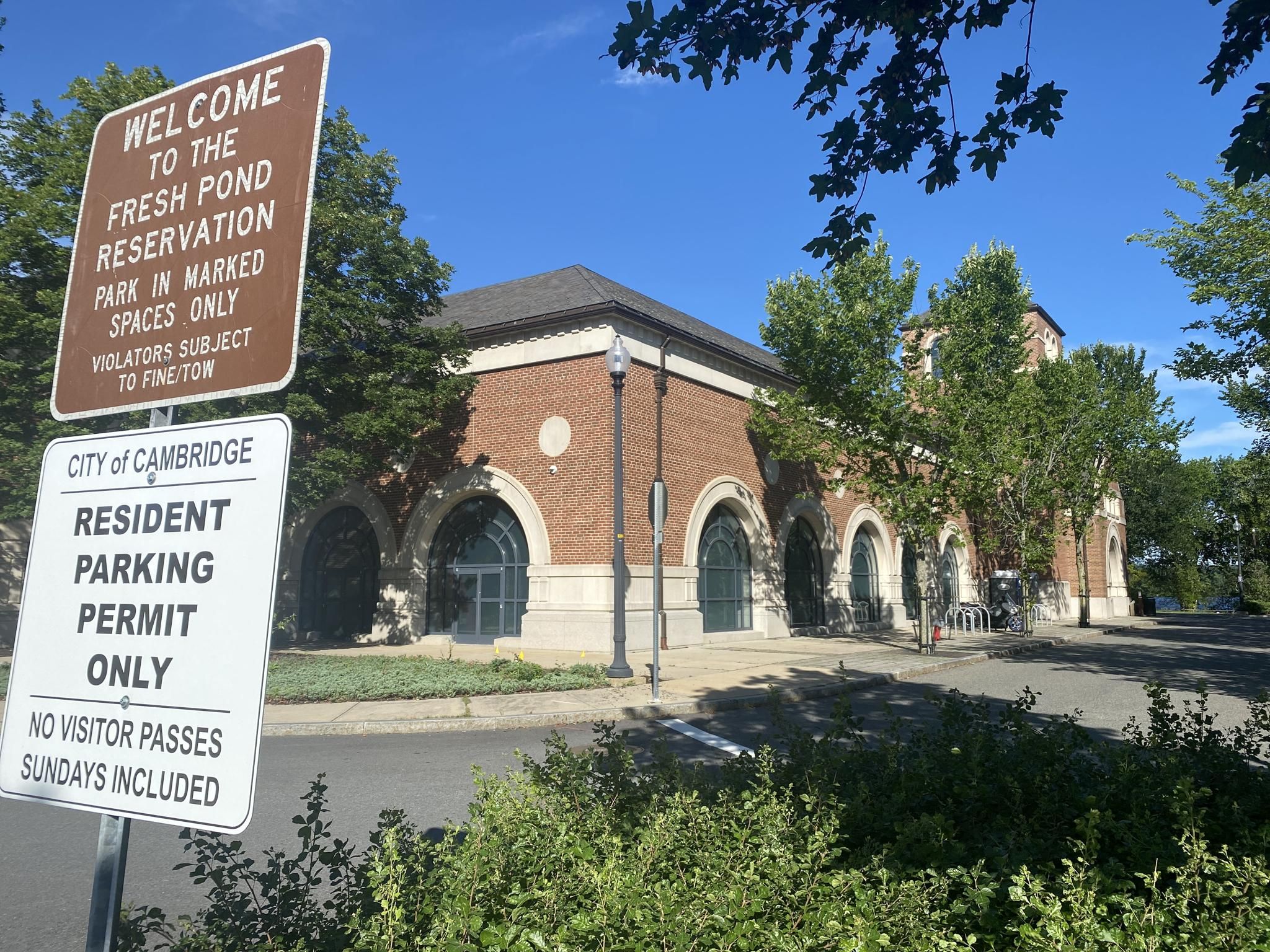 Brown brick building with arched windows, two signs, and green trees on a sunny day. - Fresh Pond Reservation Loop Trail