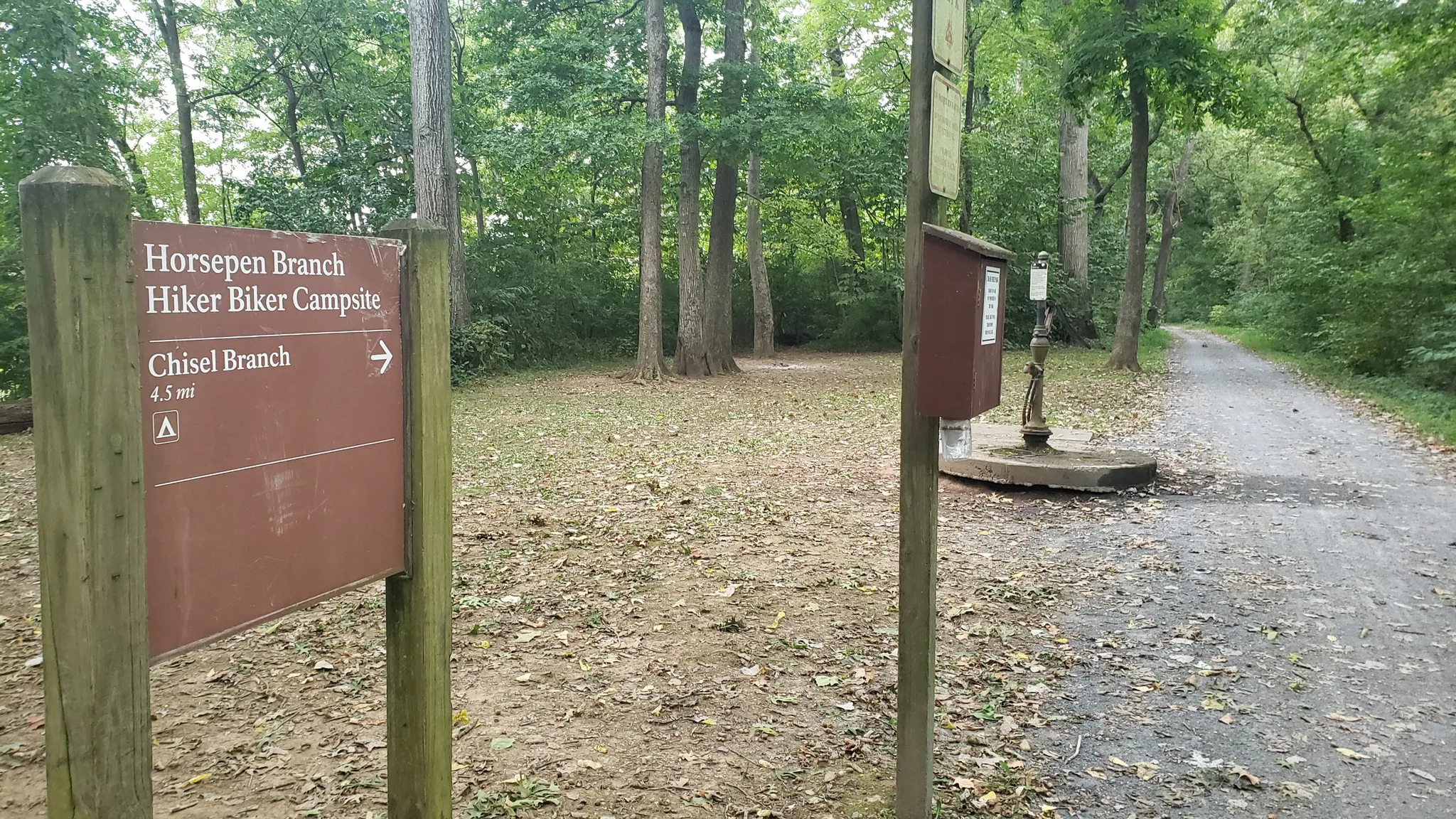 Brown signpost and water pump beside a dirt path in a shaded forest. - Chesapeake and Ohio Canal Towpath