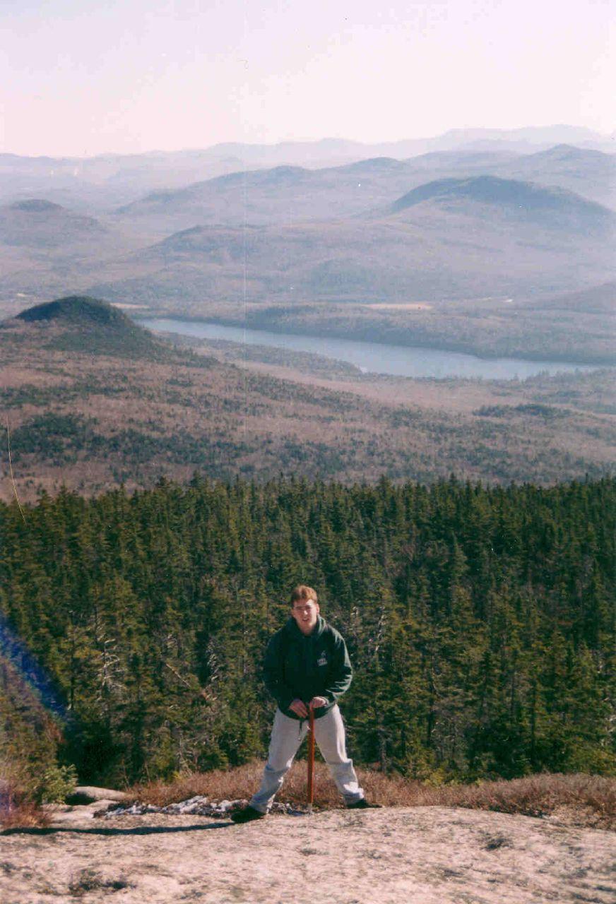 Man stands on a rocky peak overlooking vast forested mountains and a lake.