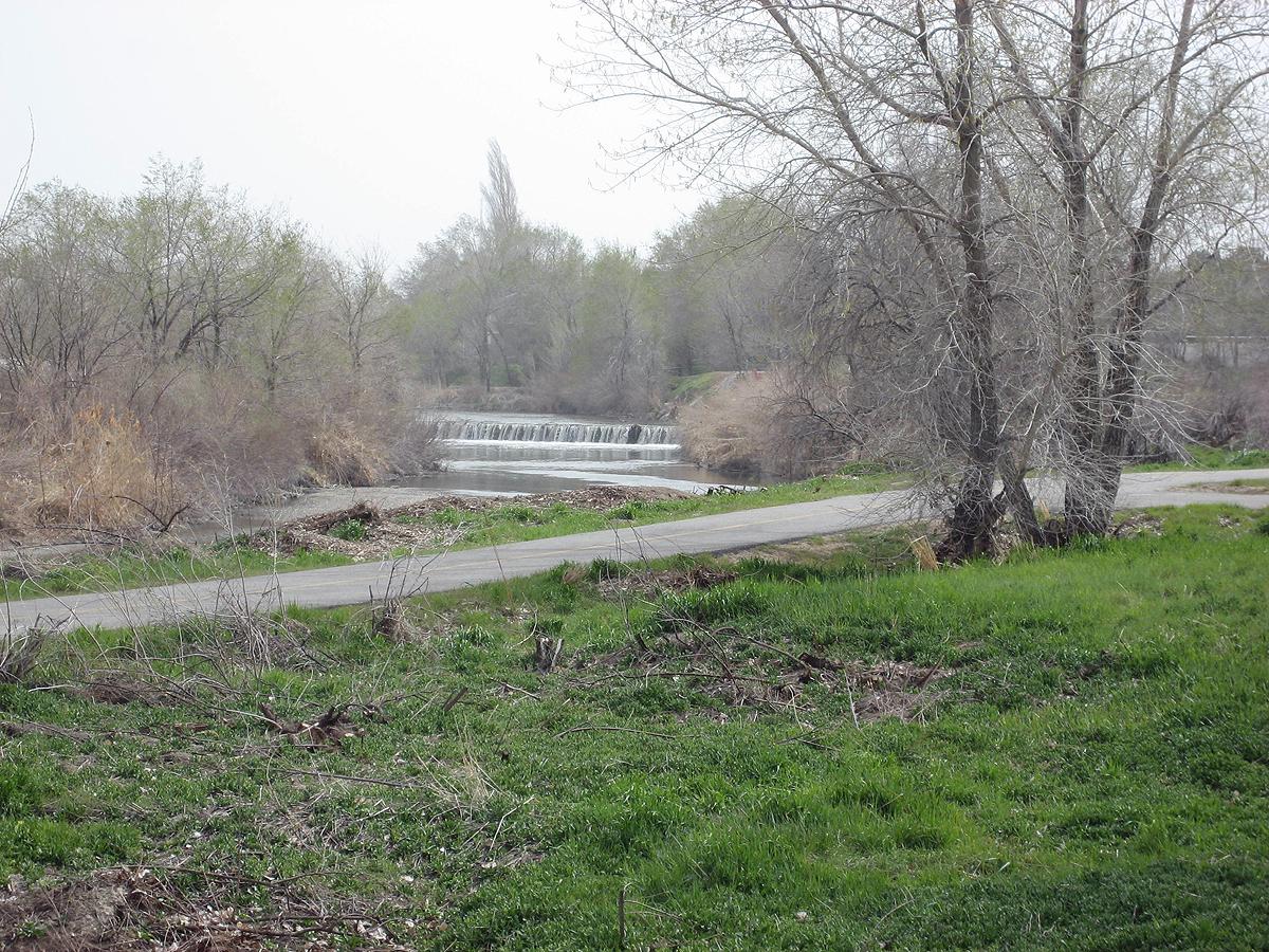 Overcast view of a river with a small dam, trees, and a paved path.