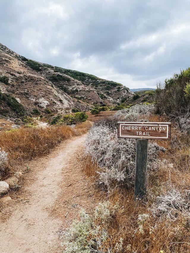 Dirt trail winding through dry hills with a trail sign under cloudy sky.
