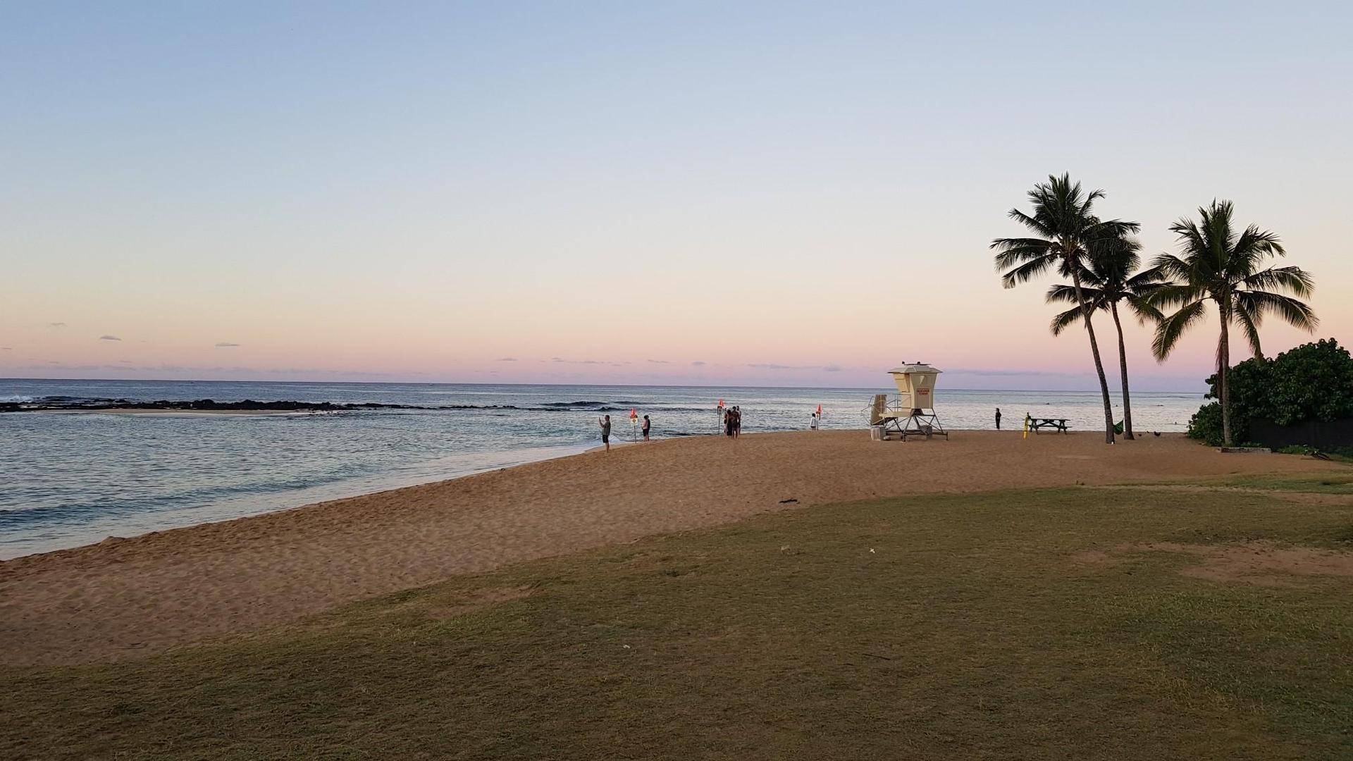 A sandy beach at dusk with a lifeguard stand, palm trees, and distant people.