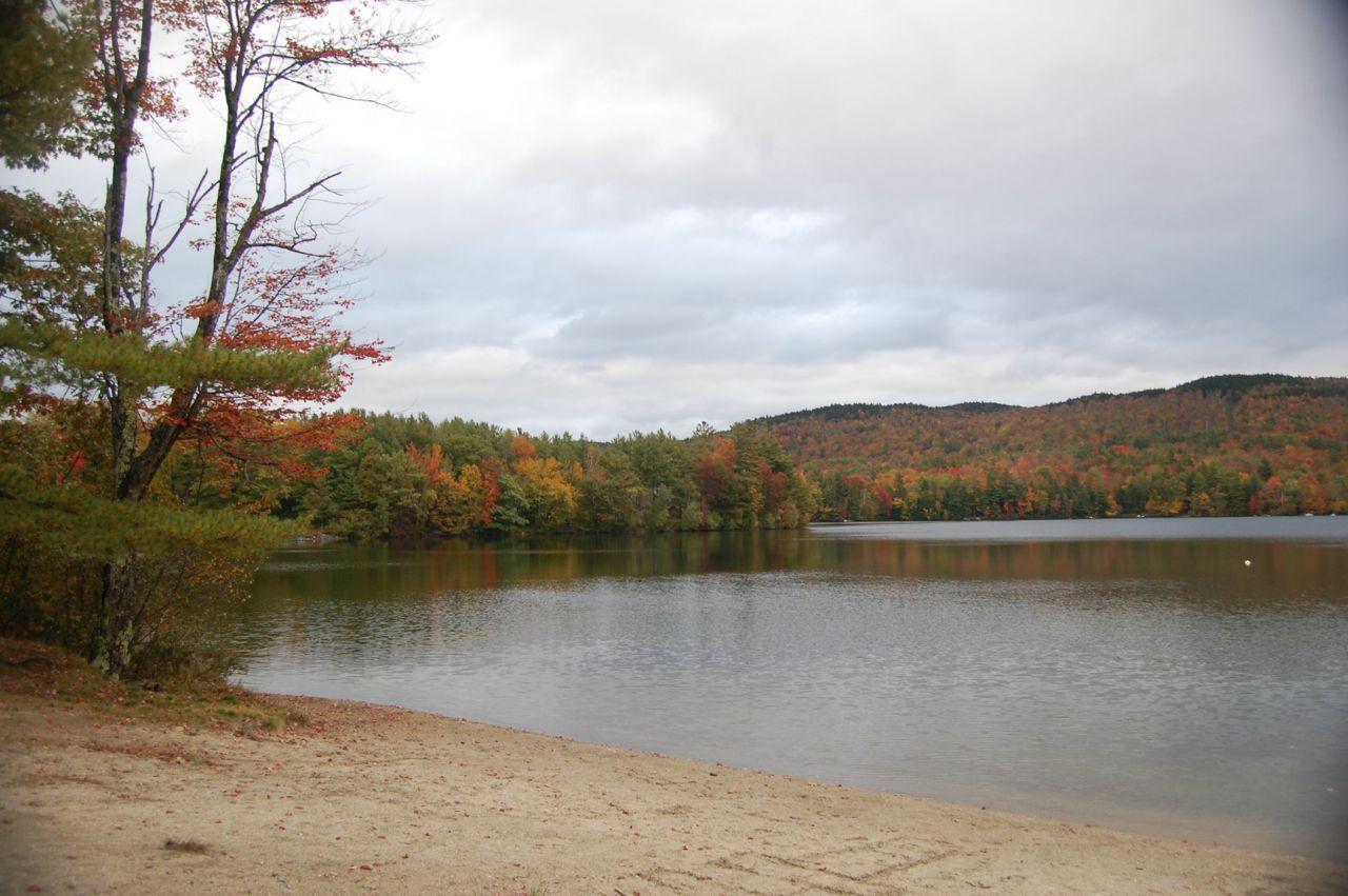 Sandy shore by a lake with autumn trees and forested hills under a cloudy sky.