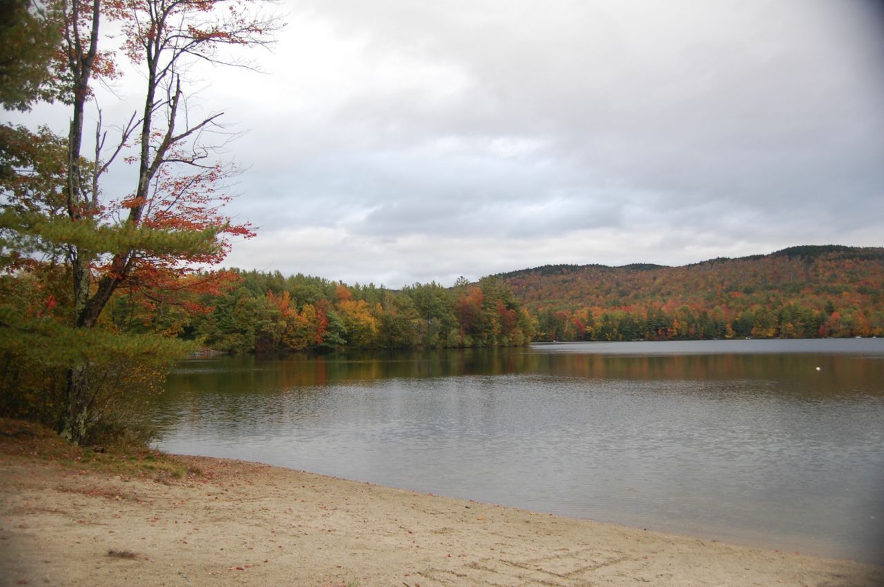 Sandy shore by a lake with autumn trees and forested hills under a cloudy sky. - Otter Pond & Lake Sunapee loop from Grantham