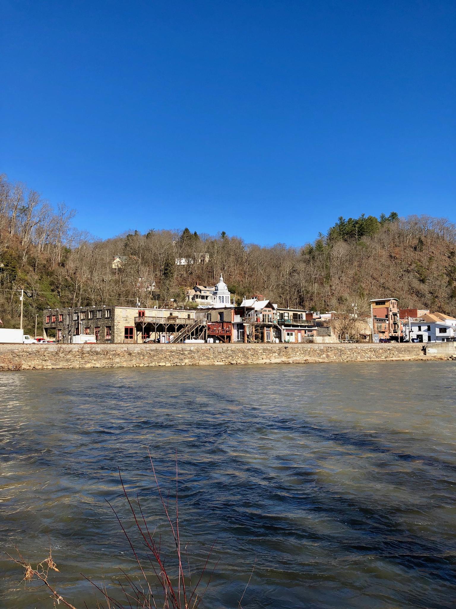 Wide river flows past buildings and a bare wooded hill on a sunny day.