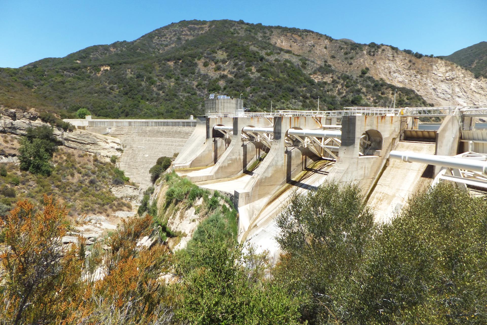 Large concrete dam structure nestled in a mountainous, arid landscape under bright sun.