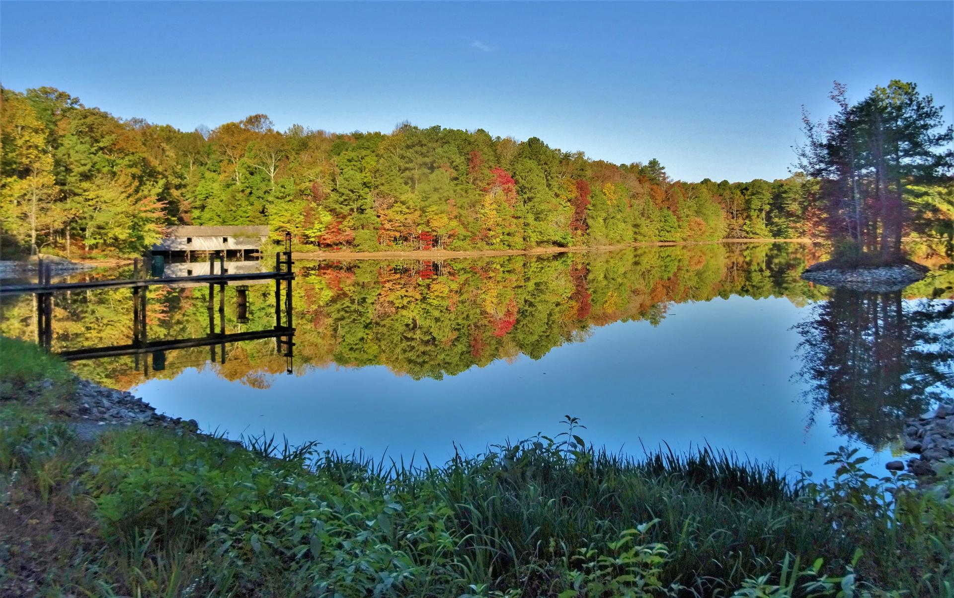 Calm lake reflecting autumn trees and a boathouse under a clear blue sky.