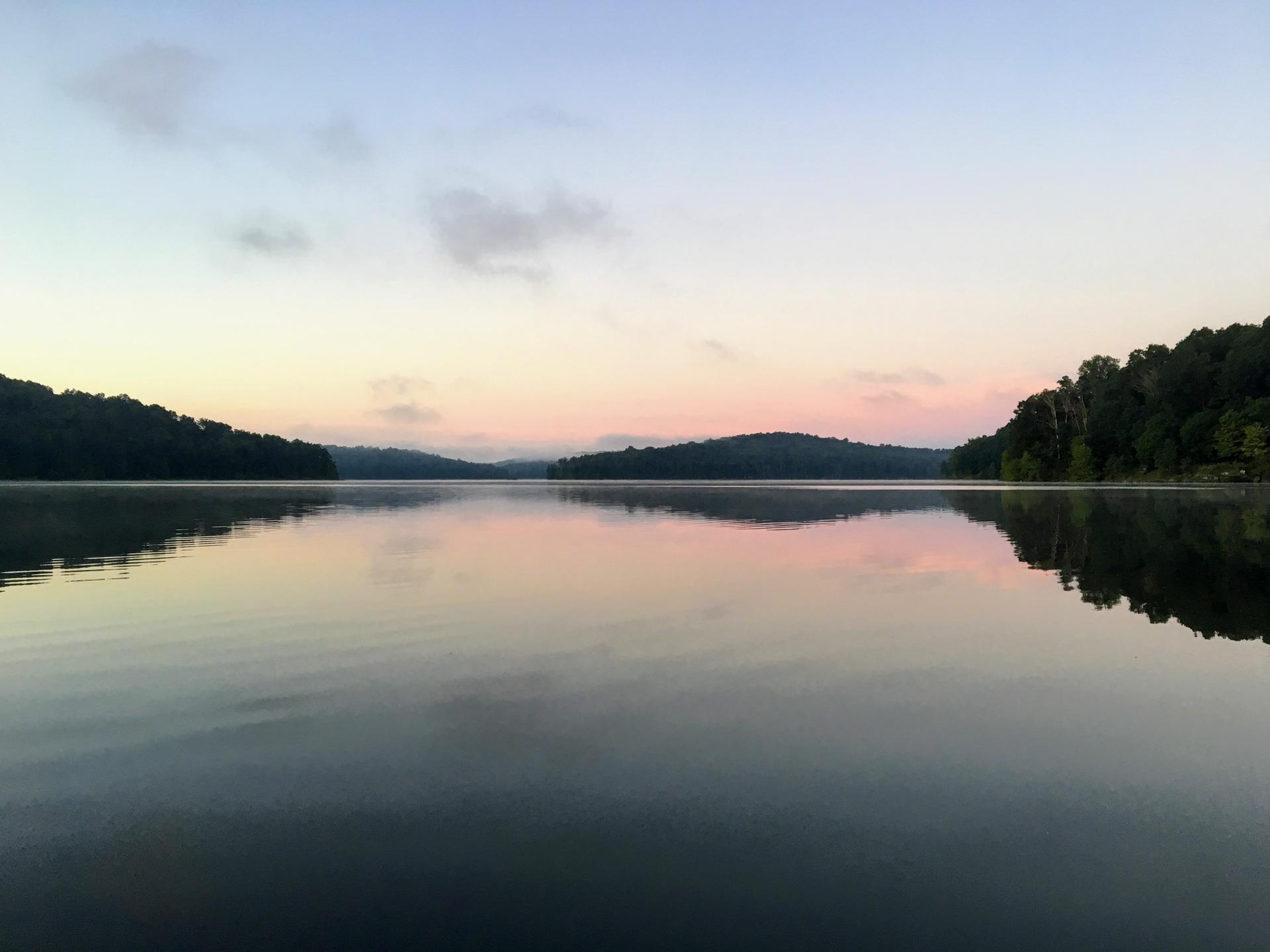 A calm lake reflects forested hills and a pastel-colored sky at dawn.